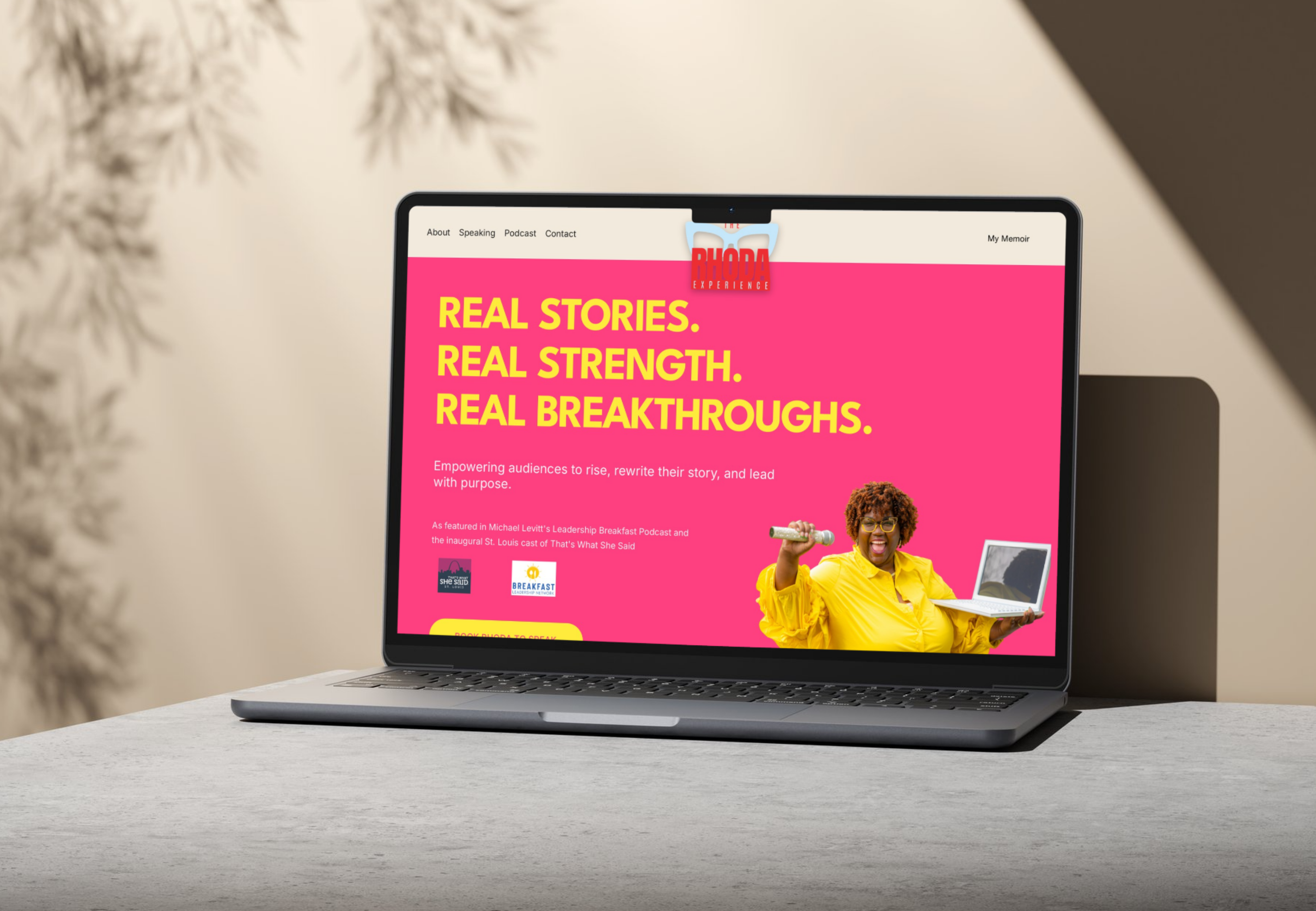 Laptop on a white desk displaying a website with bright pink background and large yellow text that reads 'Real Stories. Real Strength. Real Breakthroughs.' The webpage features a woman with curly hair, glasses, wearing a yellow shirt, holding a microphone and a laptop, smiling. The website has navigation links at the top and logos towards the bottom.