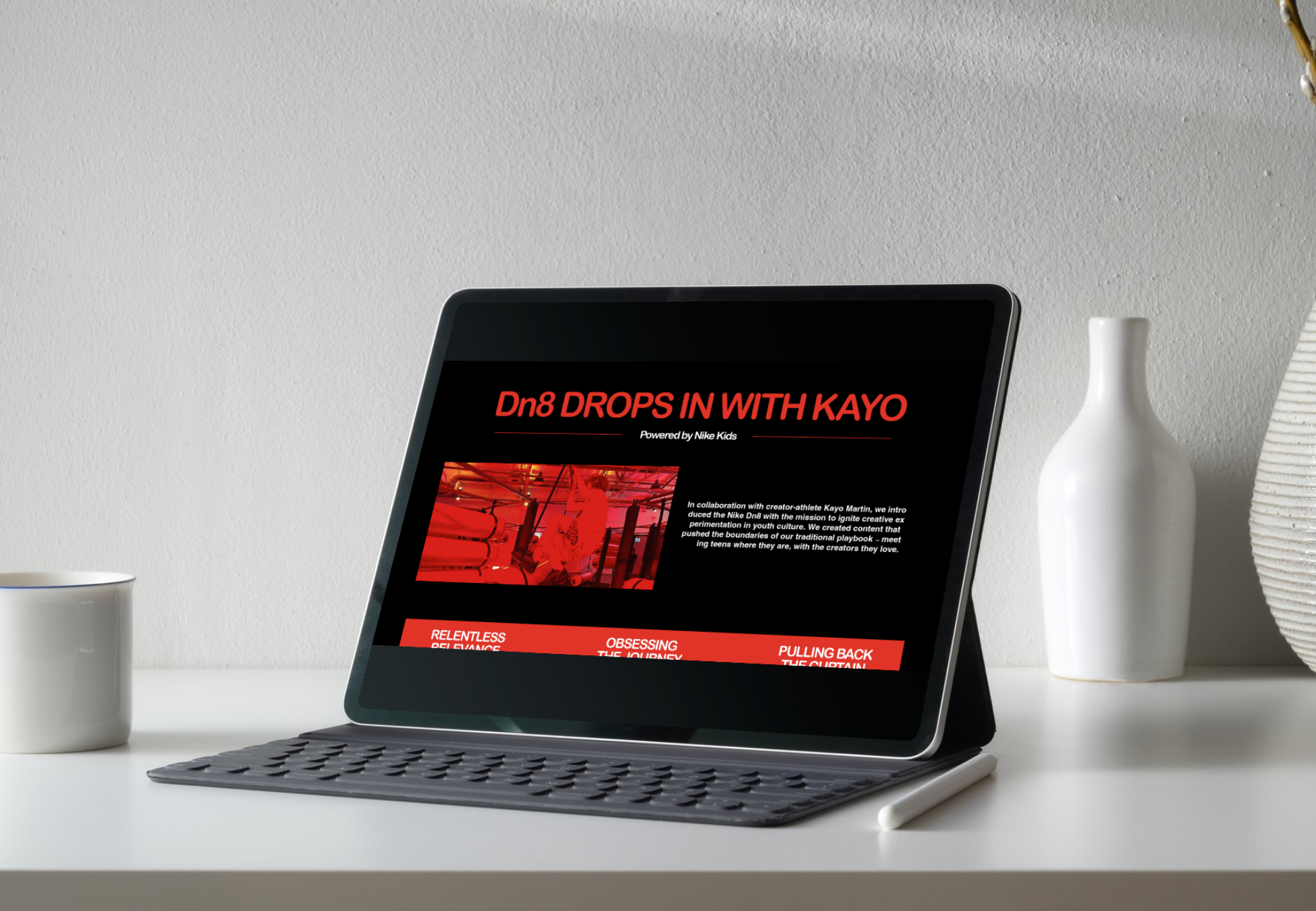 A tablet with a keyboard and stylus on a white desk displays a website titled "Dn8 DROPS IN WITH KAYO" in red and white text, with a black background and red accent sections, next to a white mug, a white vase, and a beige textured pot on the desk.