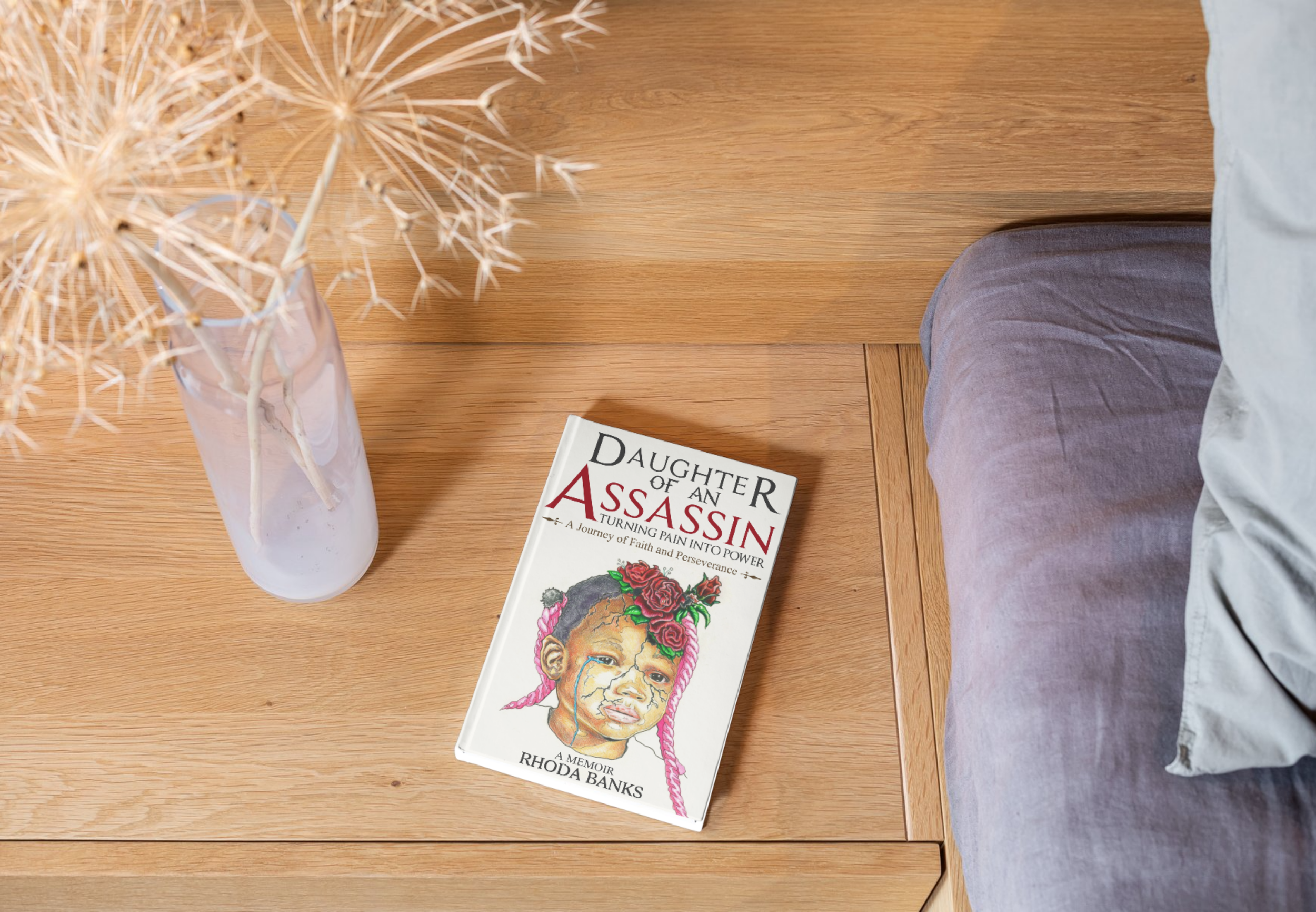 A wooden nightstand with a book titled 'Daughter of an Assassin' by Rhoda Banks, a vase with dried flowers, and part of a bed with gray bedding.