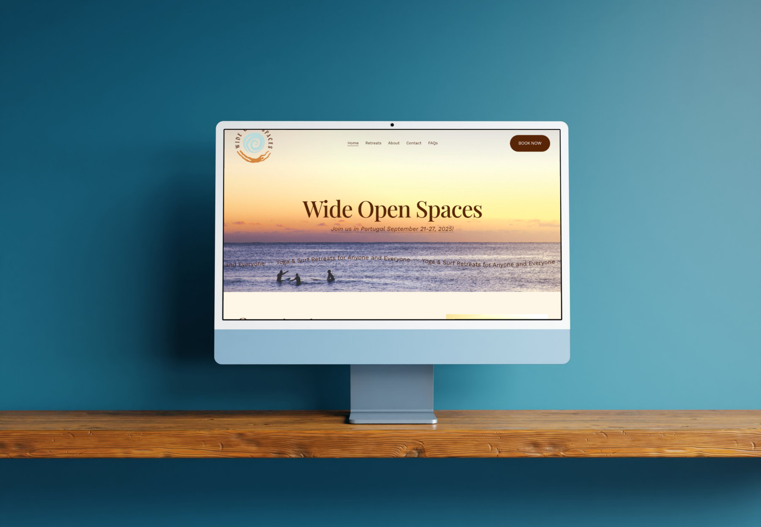 Computer monitor on a wooden shelf displaying a website for Wide Open Spaces with a sunset over the ocean and three silhouetted surfers in the water.