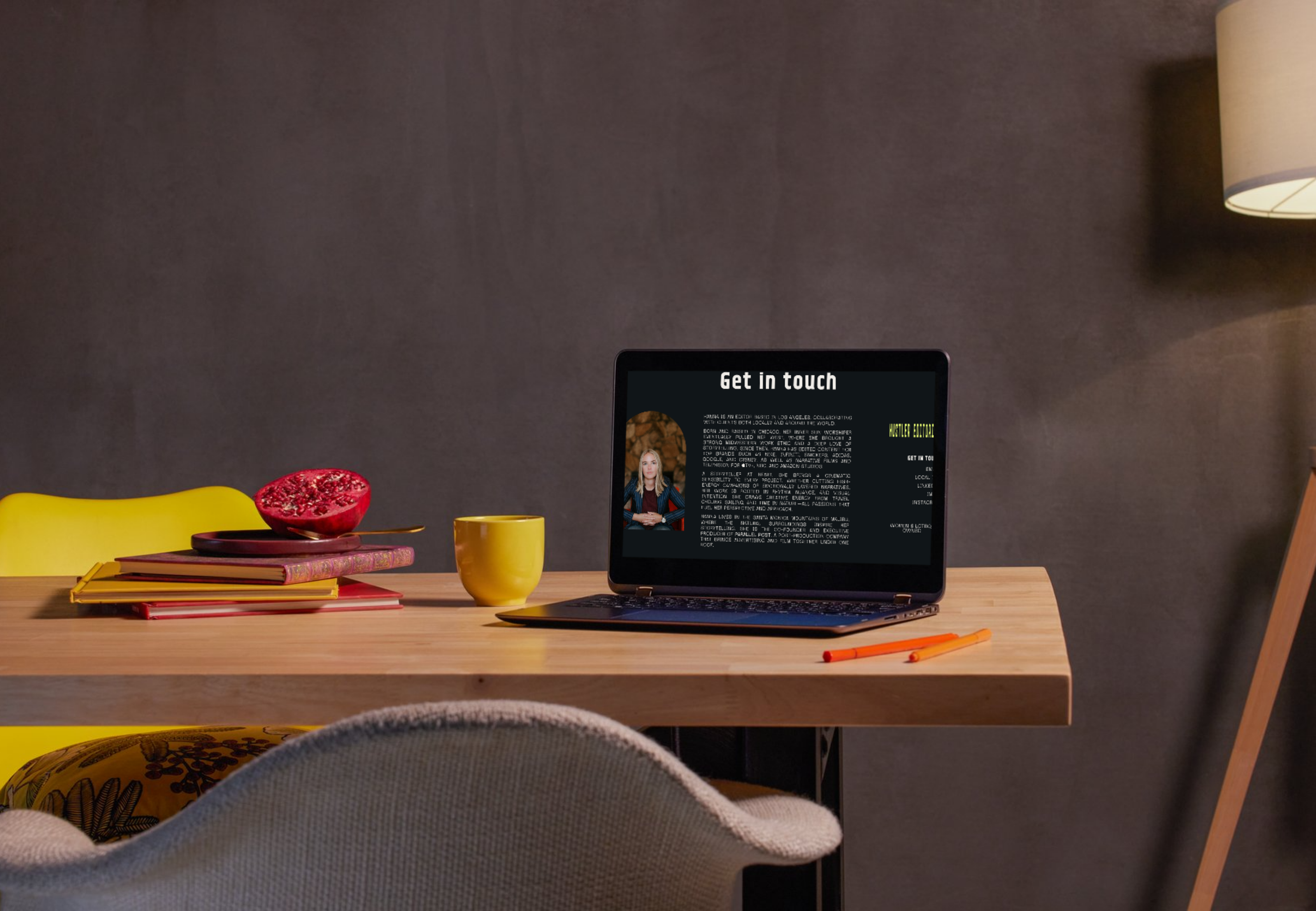A wooden desk with yellow chair, yellow mug, books, half-cut pomegranate, and orange pens. A laptop displays a 'Get in touch' webpage. The background wall is gray, and there is a lamp on the right.