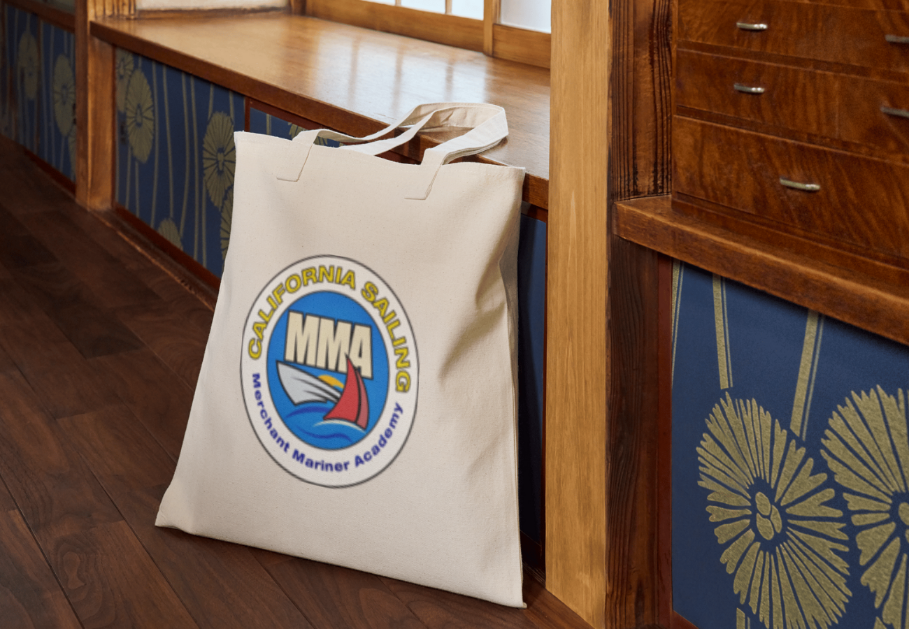 A white tote bag with the MMA logo and 'California Sailing' text leans against a wooden wall in a room with wooden floors and patterned blue and gold decor.