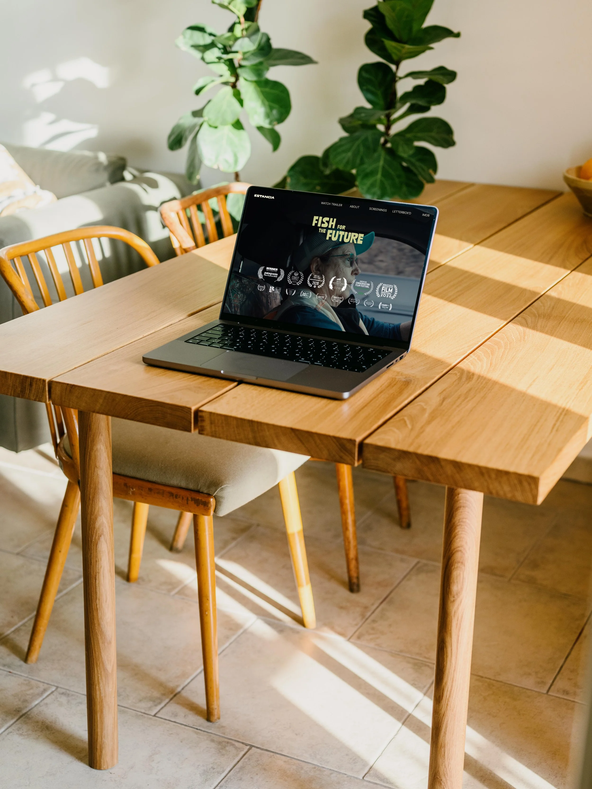 A wooden dining table with a MacBook laptop open displaying a movie or documentary titled 'Fish for the Future,' with greenery in the background and sunlight casting shadows on the tiles and table.