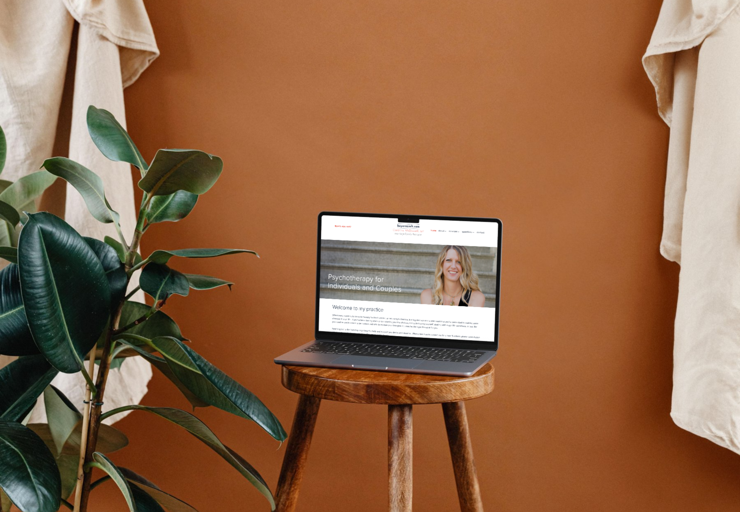 Laptop on wooden stool displaying a psychotherapy website with a smiling woman on the screen, next to a green leafy plant, against a brown wall with cream-colored curtains.