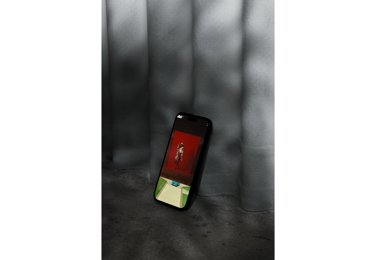 A smartphone resting against a textured gray curtain on a concrete floor, displaying an image of a person standing in front of a red backdrop.