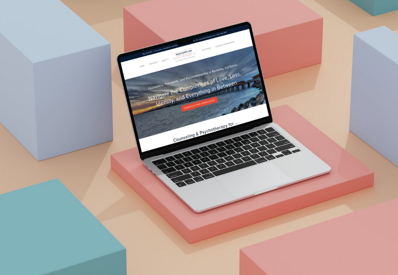 A MacBook laptop displaying a therapy website, placed on pink and peach-colored geometric blocks, with additional pastel blue blocks around.