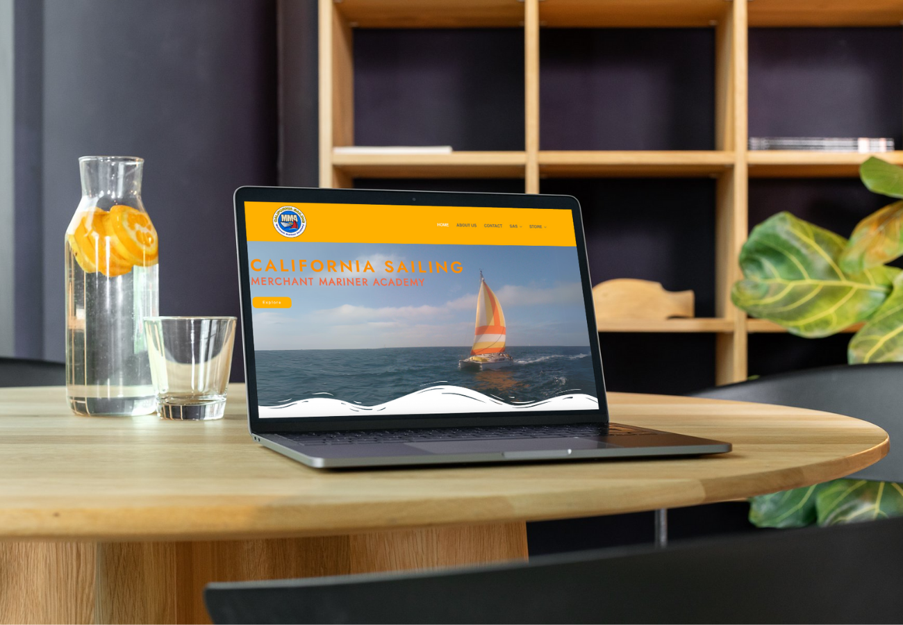 Desk with a laptop displaying a website for California Sailing, a sailing academy, with a picture of a sailboat on the ocean. There is a glass of water with orange slices and a pitcher of water on the left, and some green plants on the right. A wooden shelf with a few objects is in the background.
