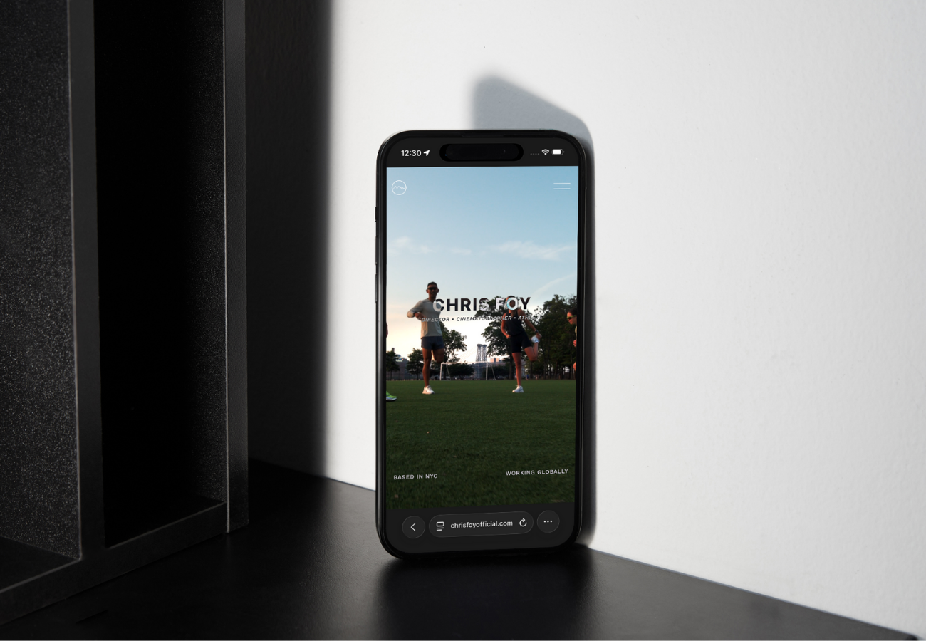 A smartphone displaying the homepage of Chris Foy's official website, standing on a black surface next to a dark textured object, with a white wall in the background. The website features a photo of two people on a grassy field under a blue sky.