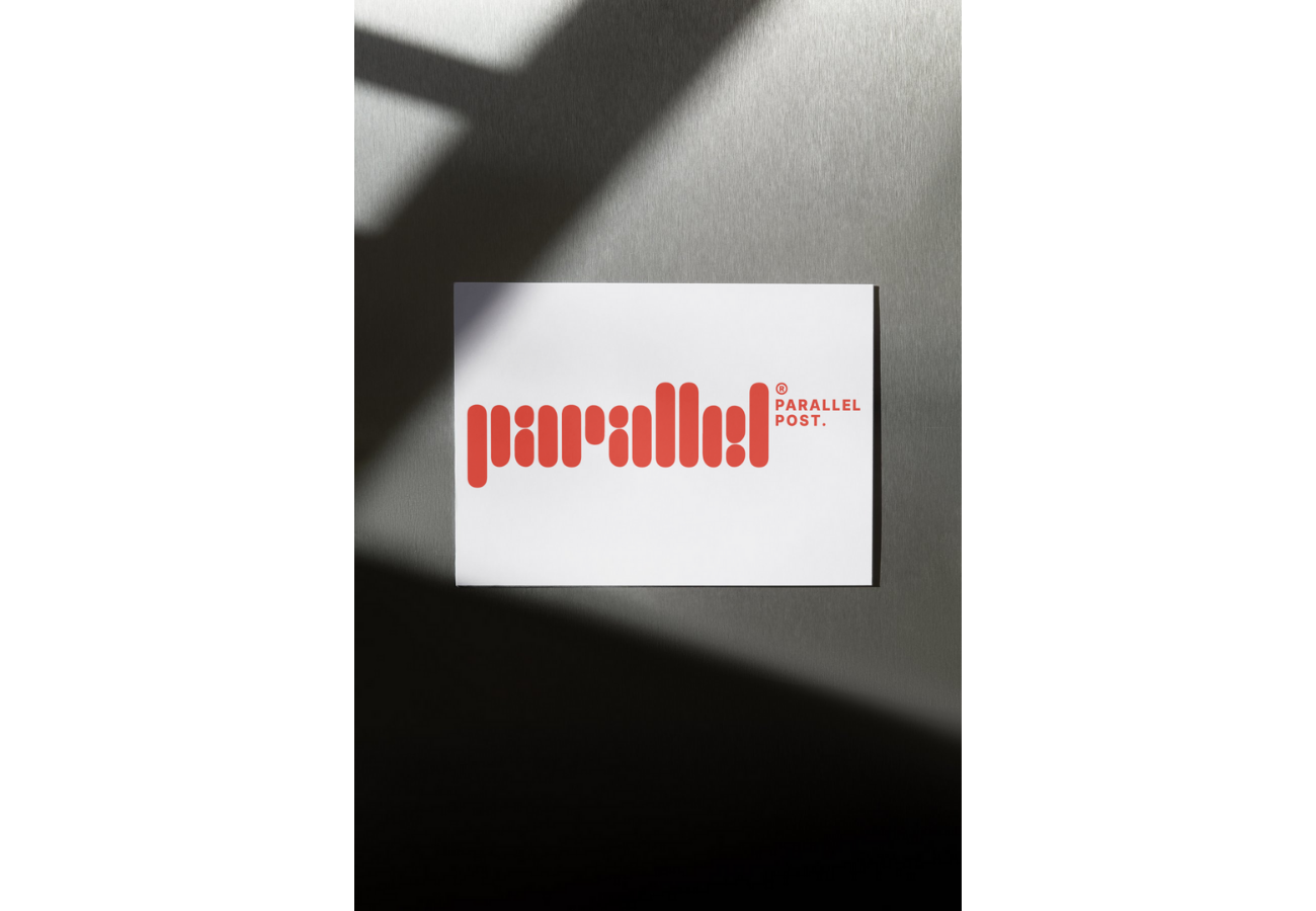 A white card with red stylized text spelling 'parallel' and smaller text 'PARALLEL POST.' placed on a gray surface with shadows crossing over it.