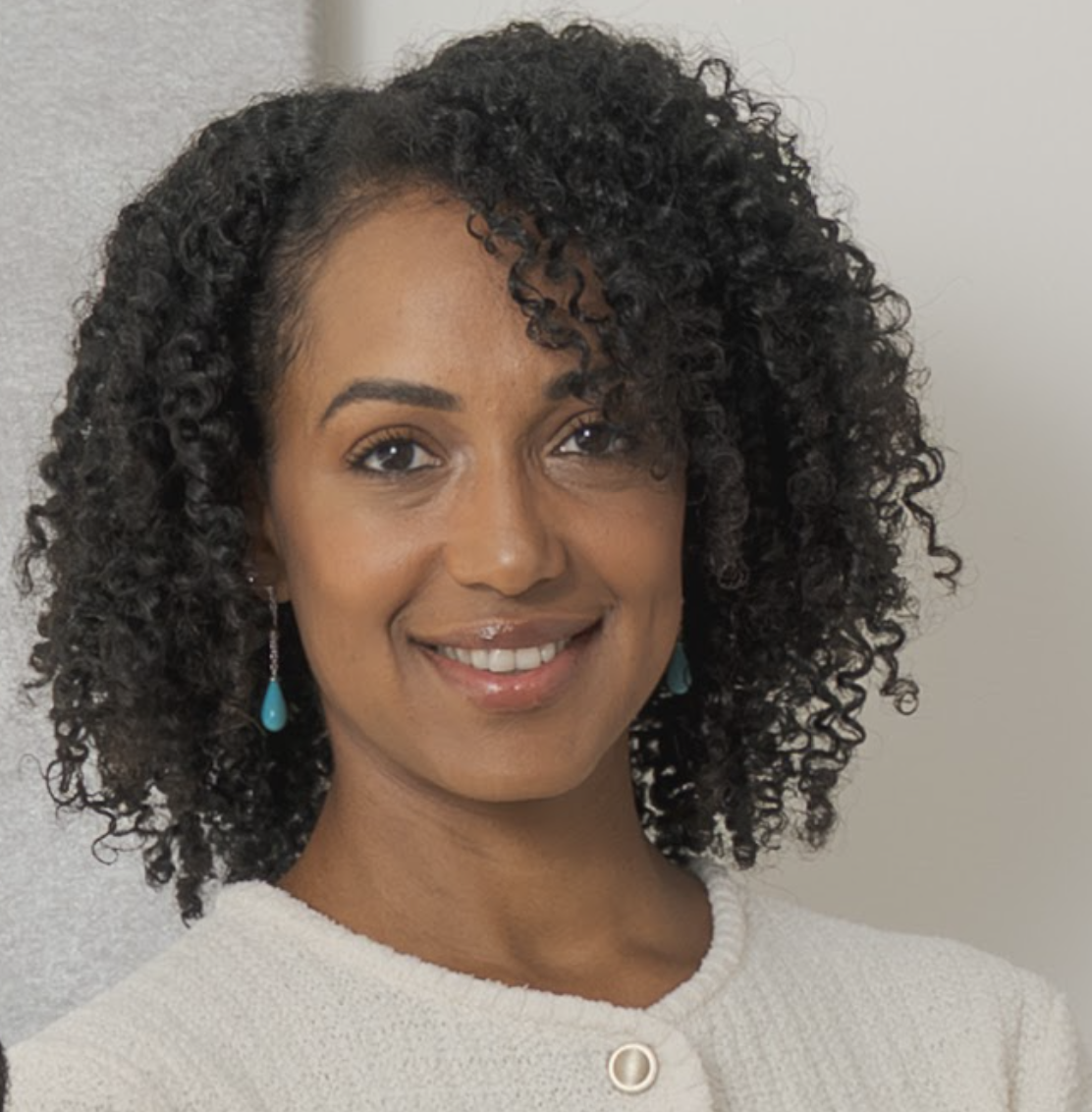 Portrait of a smiling woman with curly black hair, wearing turquoise earrings and a white textured top, standing against a neutral background.