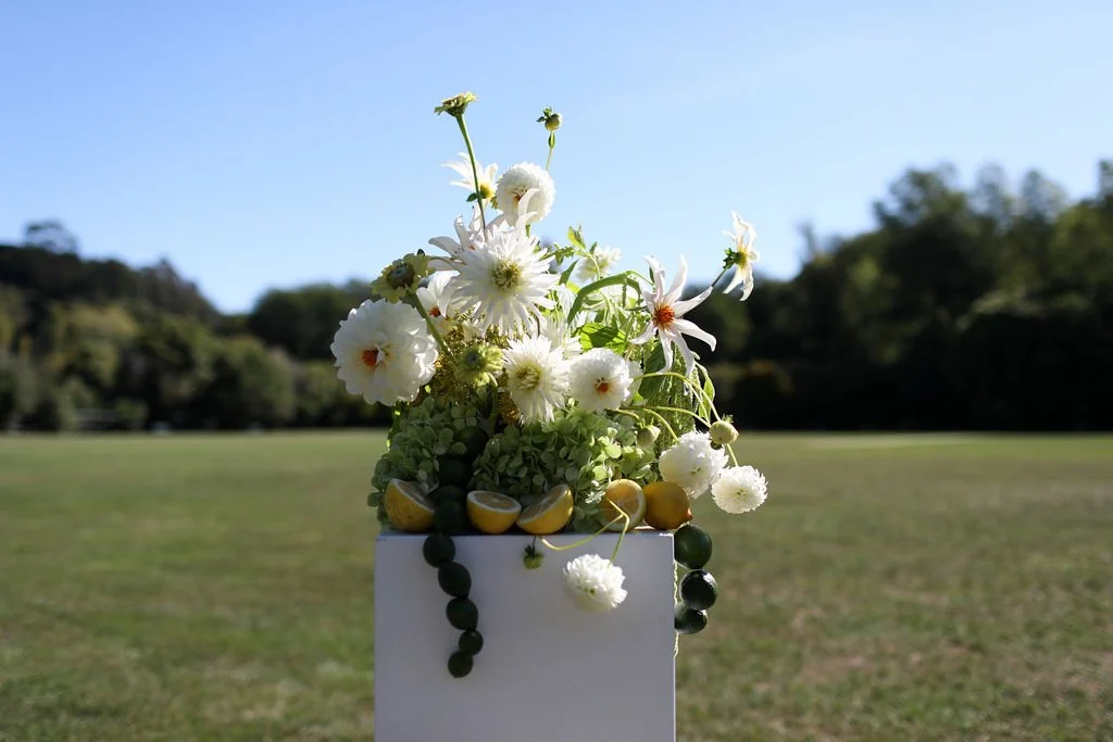 A bouquet of white flowers in a white vase outdoors on a grassy field with trees and a clear blue sky in the background.