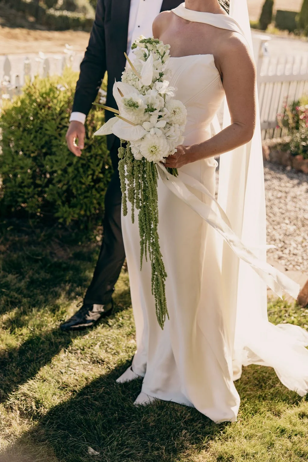 Bride in a white dress holding a cascading white floral bouquet during outdoor wedding ceremony.