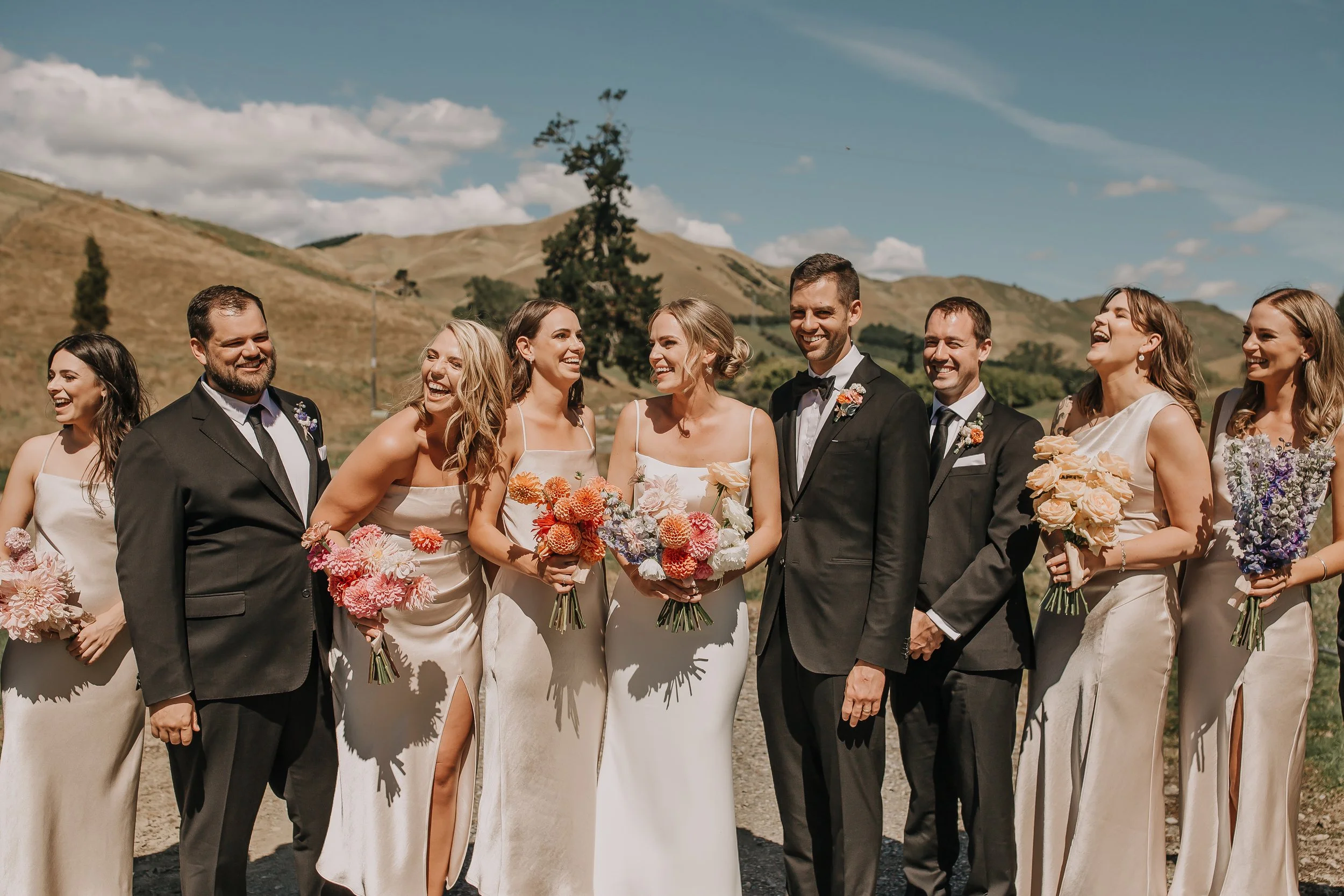 A wedding party posing outdoors with mountains in the background. The group includes men in suits and women in cream dresses, some holding bouquets. Everyone is smiling and laughing.