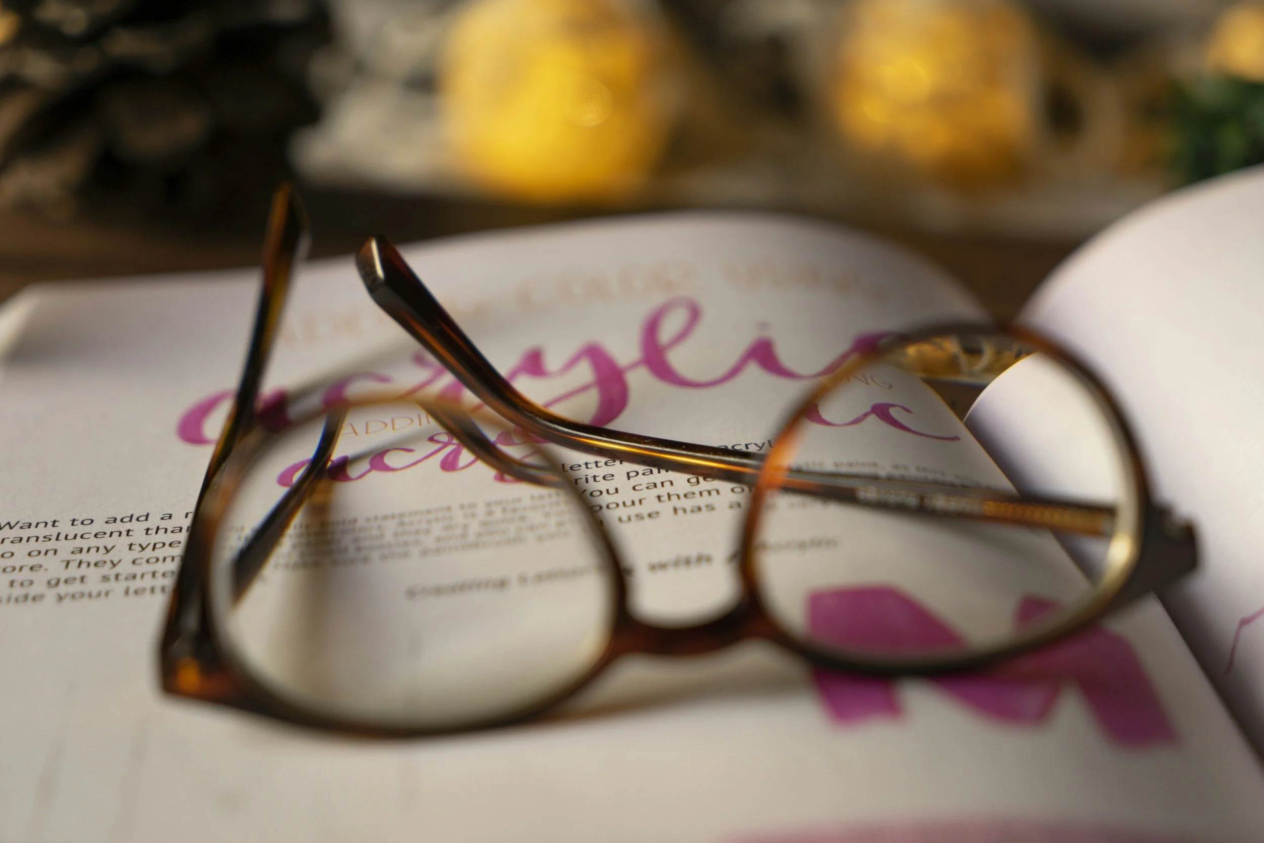 Close-up of a pair of glasses resting on a paper with pink and black text, blurry yellow objects in the background.