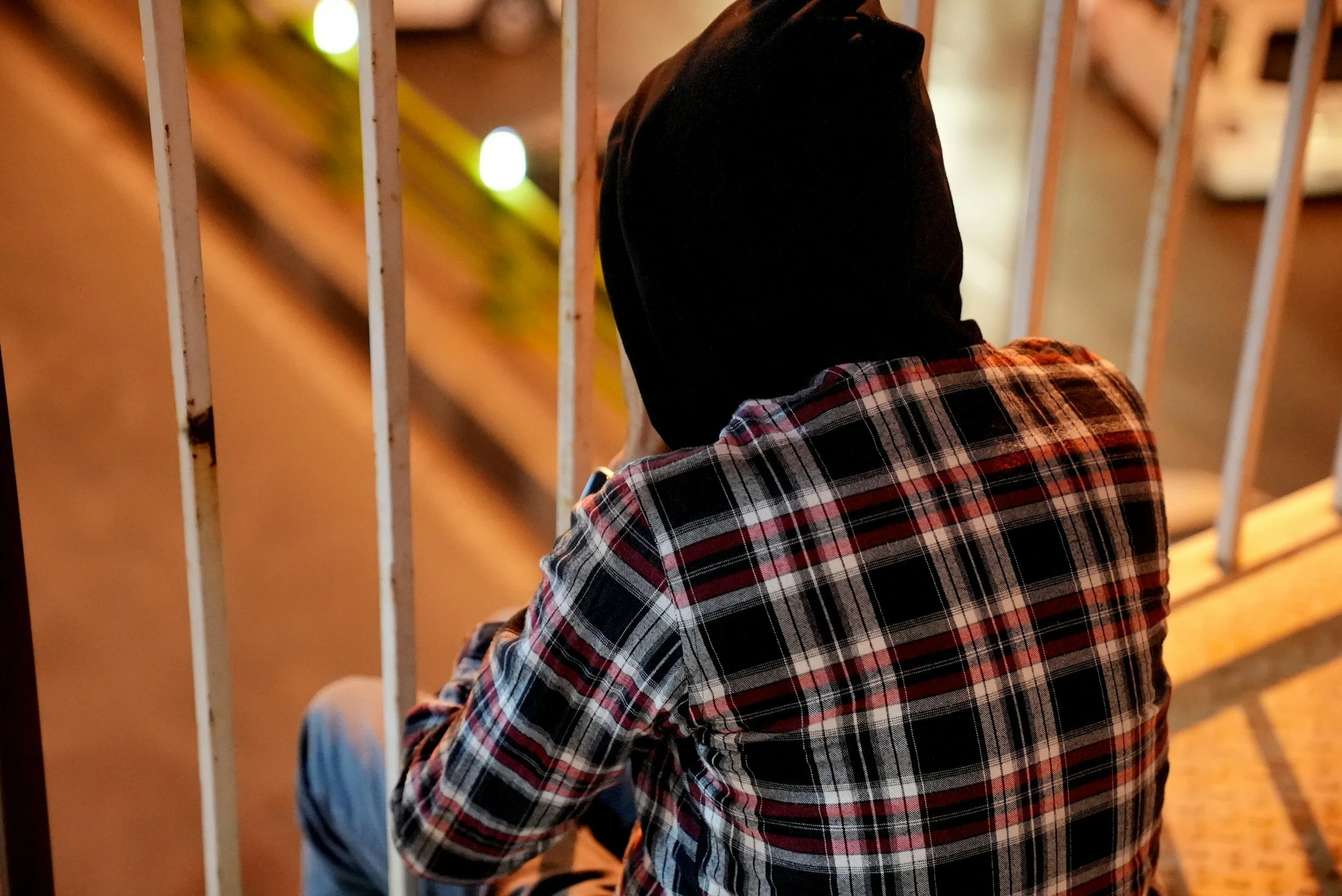 Unacceptable behavior of a young person sitting behind metal bars at night, wearing a black hoodie and a plaid shirt.