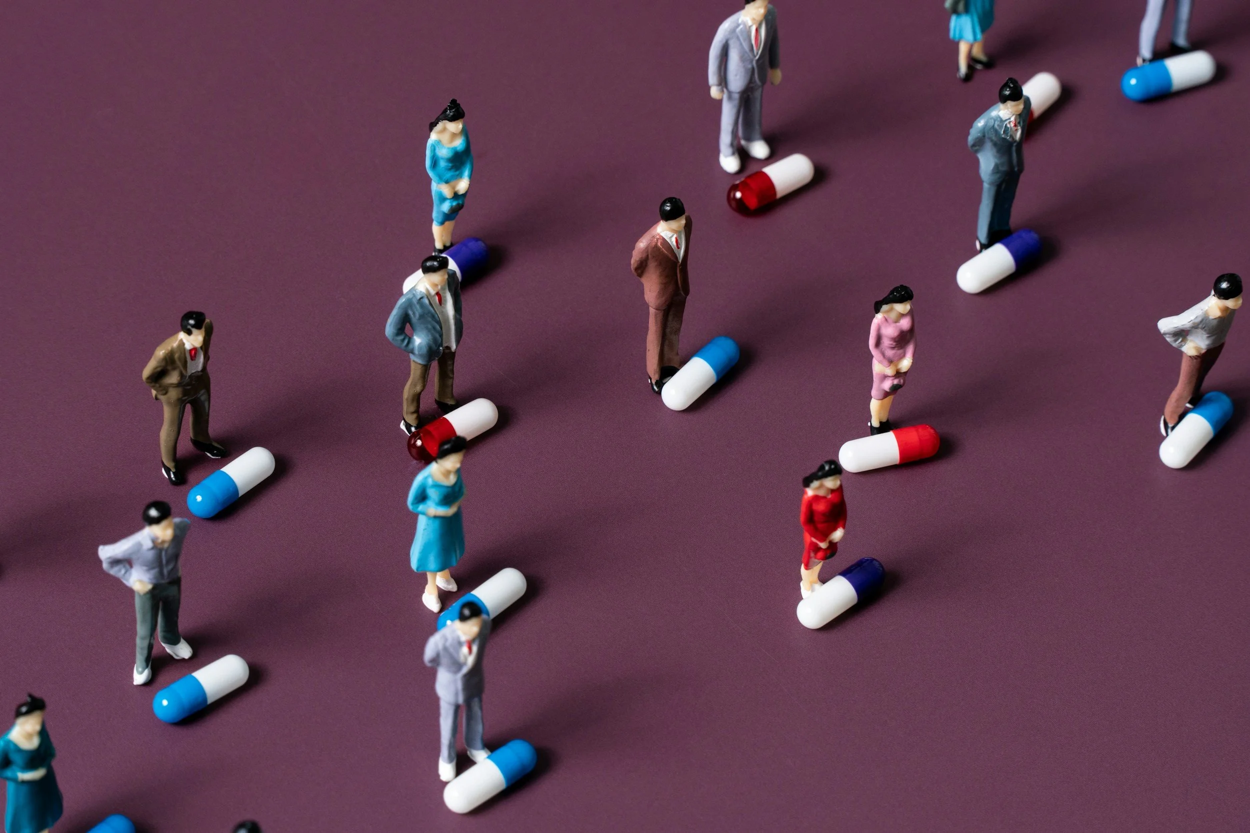 Pill shaming - Small figurines of people standing on capsules arranged on a purple surface.