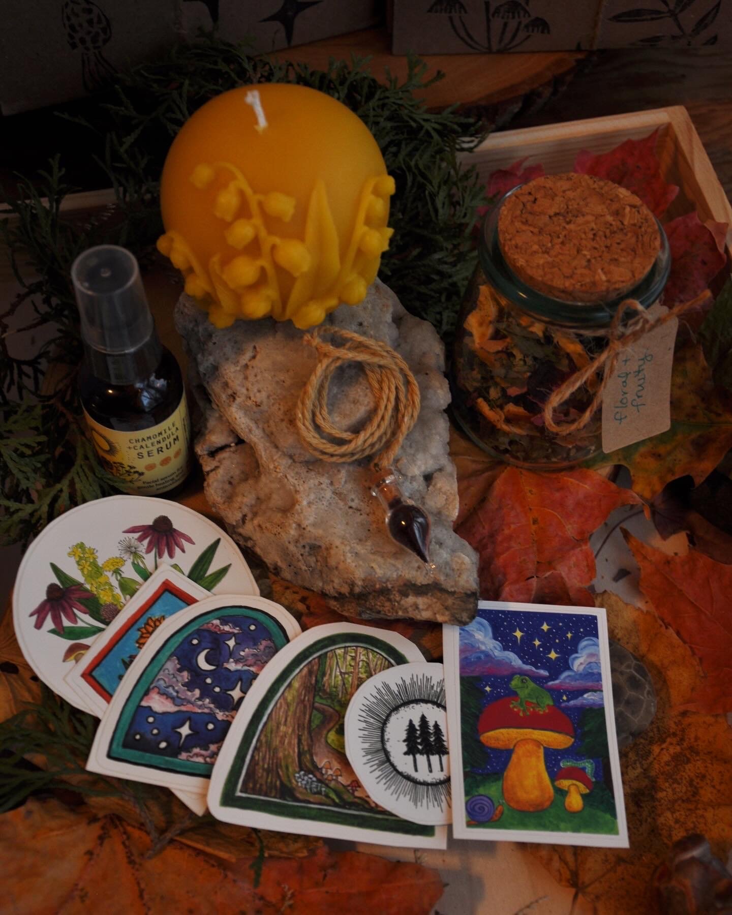 A collection of spiritual and herbal items, including a yellow decorated candle, a bottle of essential oil, a jar of dried herbs labeled 'flower + fruit,' several colorful oracle cards, and natural elements like leaves and rocks, arranged on a wooden