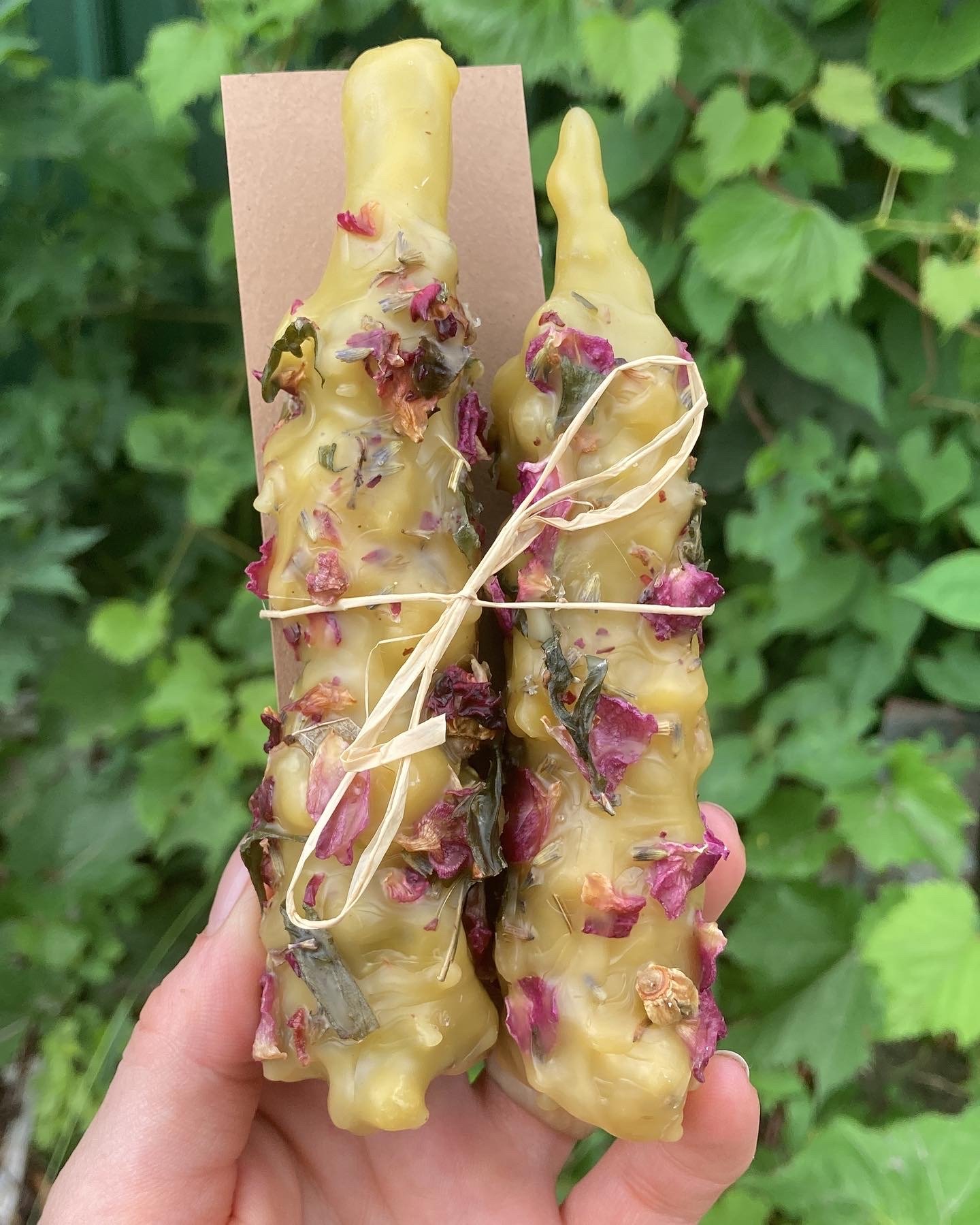 Hand holding two large beeswax candles with purple, pink, and green flower petals embedded in the wax, tied with a thin string, against a background of green leaves.
