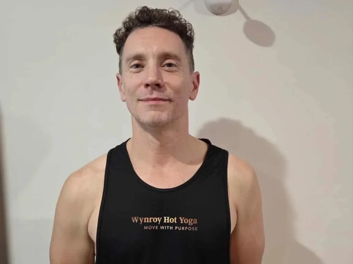 Man wearing a black tank top with 'Wynroy Hot Yoga, Move with Purpose' written on it, standing in front of a plain wall.