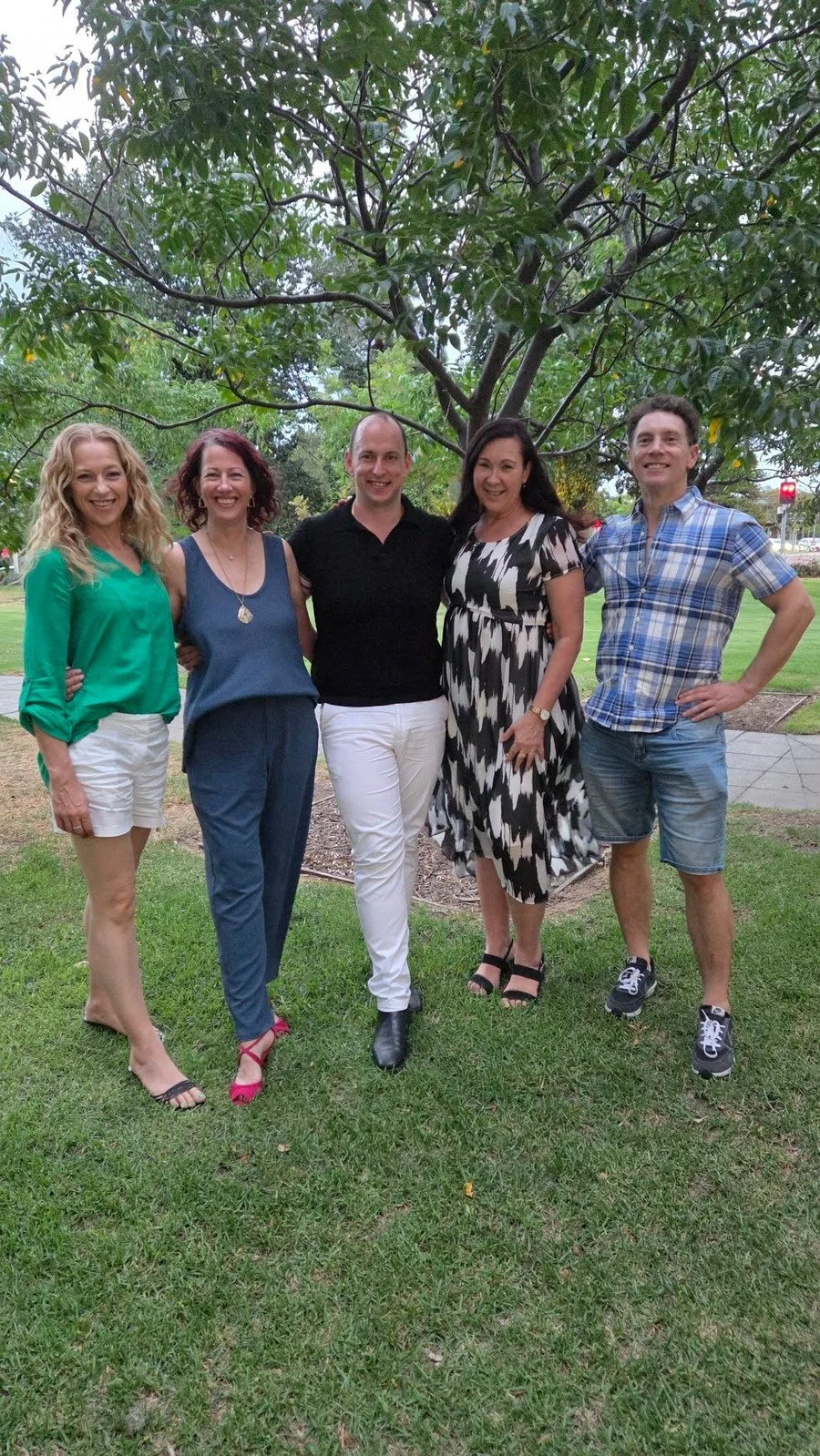 Group of five people standing outdoors on grass, smiling, with a large tree in the background.