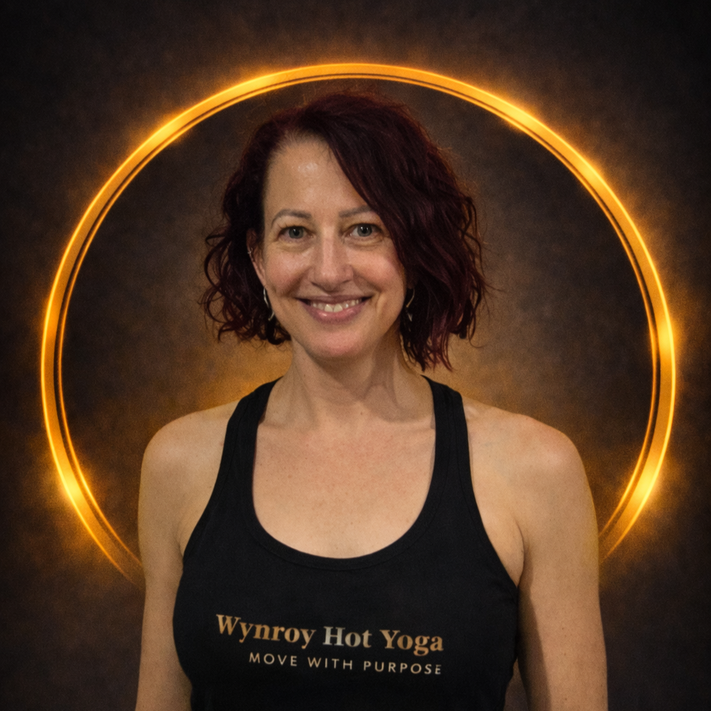 Smiling woman with red curly hair wearing a black tank top with 'Wynroy Hot Yoga' logo, standing in front of a glowing circular light on a dark background.