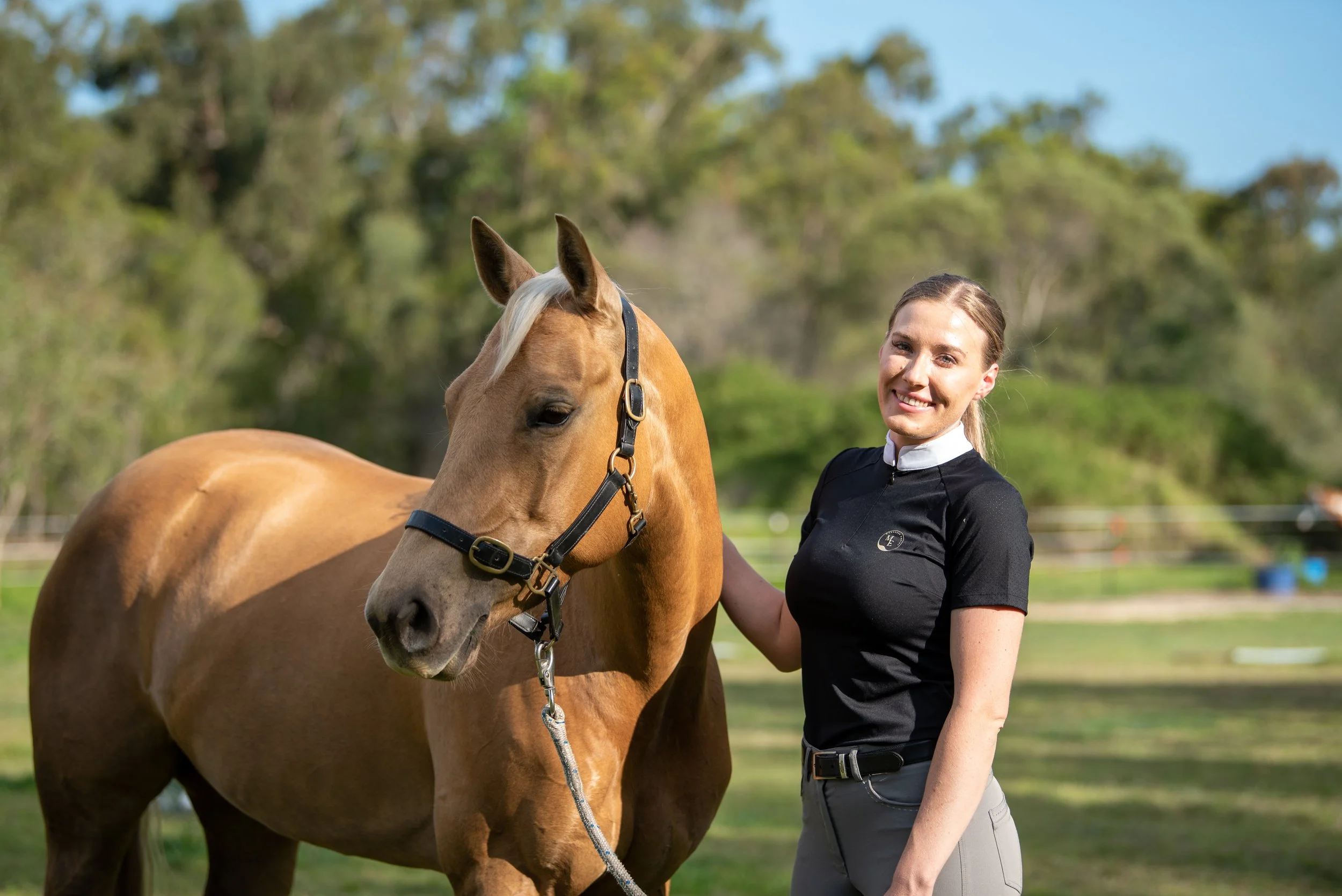A smiling woman in a black riding shirt standing next to a light brown horse with a blonde mane outdoors with trees in the background.