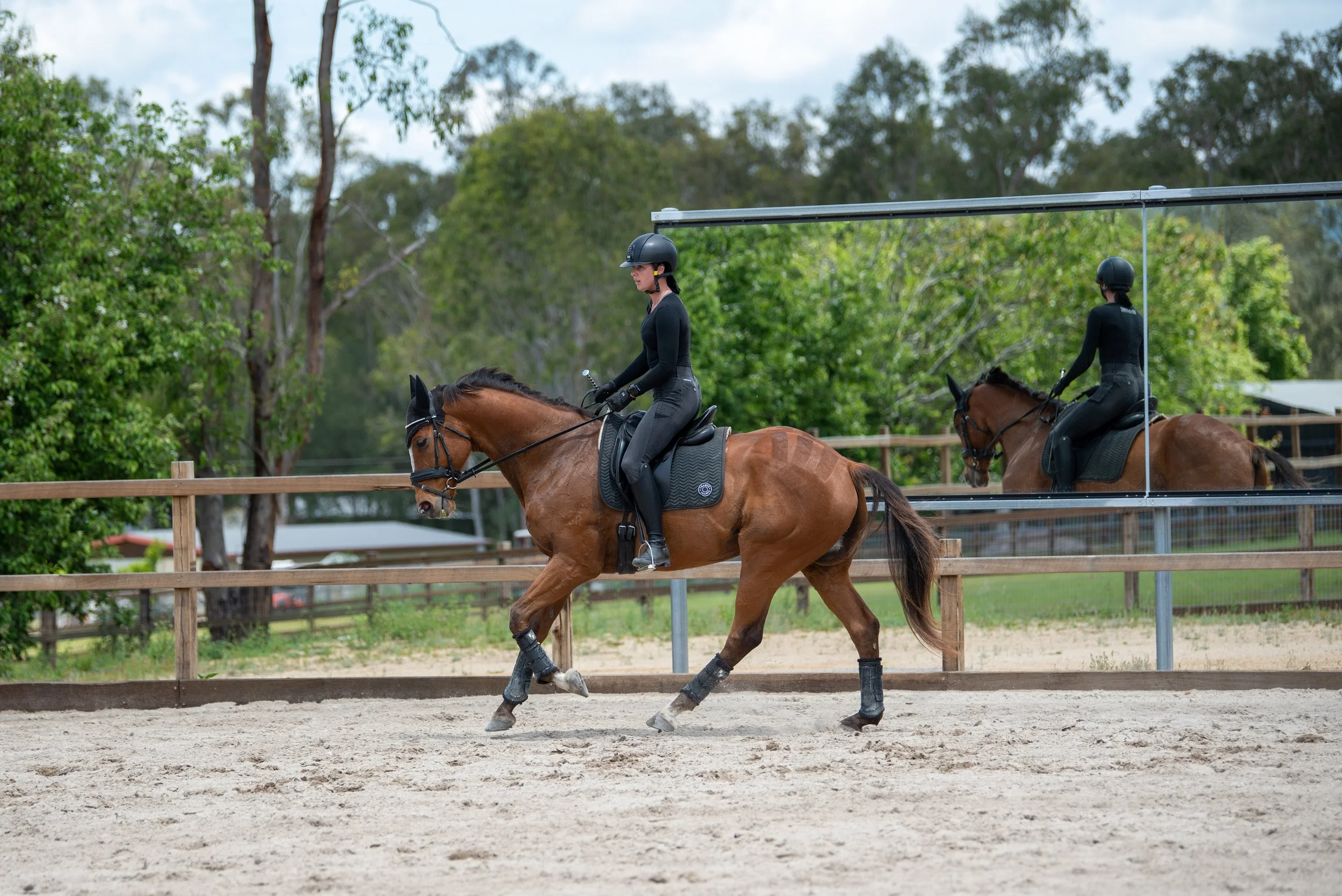 A person riding a horse in an outdoor riding arena with trees and a mirror in the background.