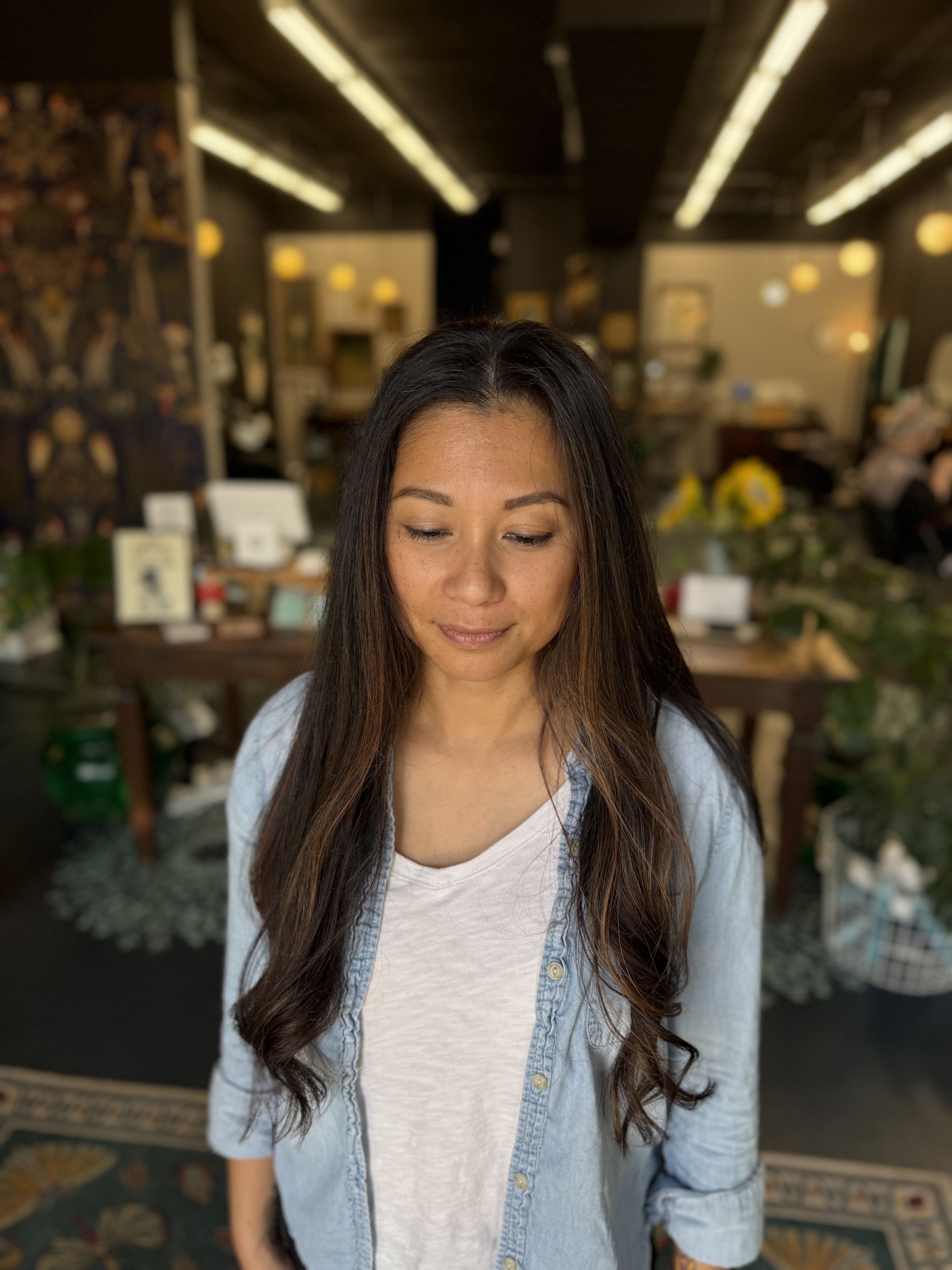 A woman with long wavy hair standing in a room with dim lighting and modern decor, wearing a white top and light denim shirt.