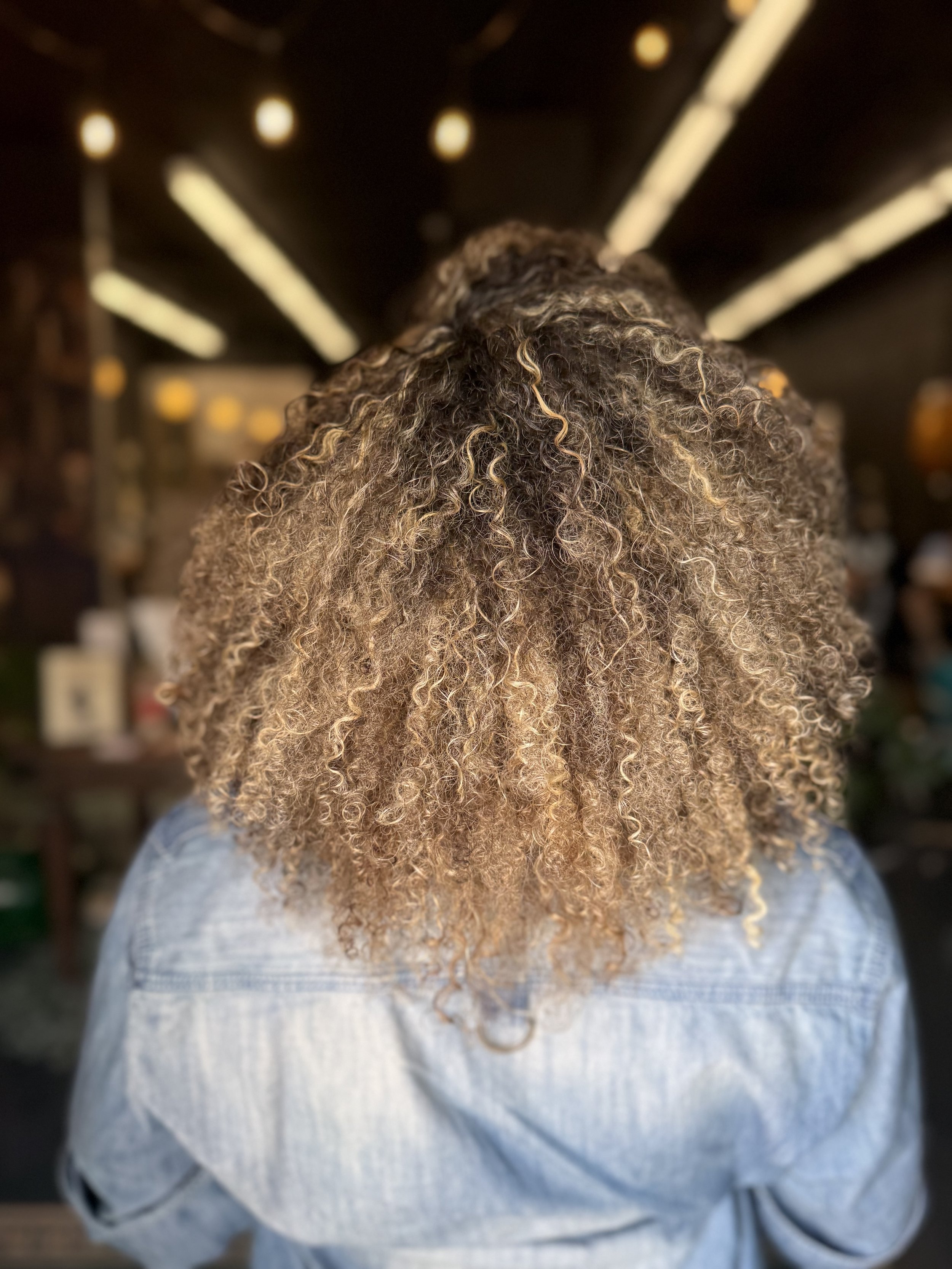 Woman with curly brown and blonde hair in a denim jacket, viewed from behind, in a dimly lit room.