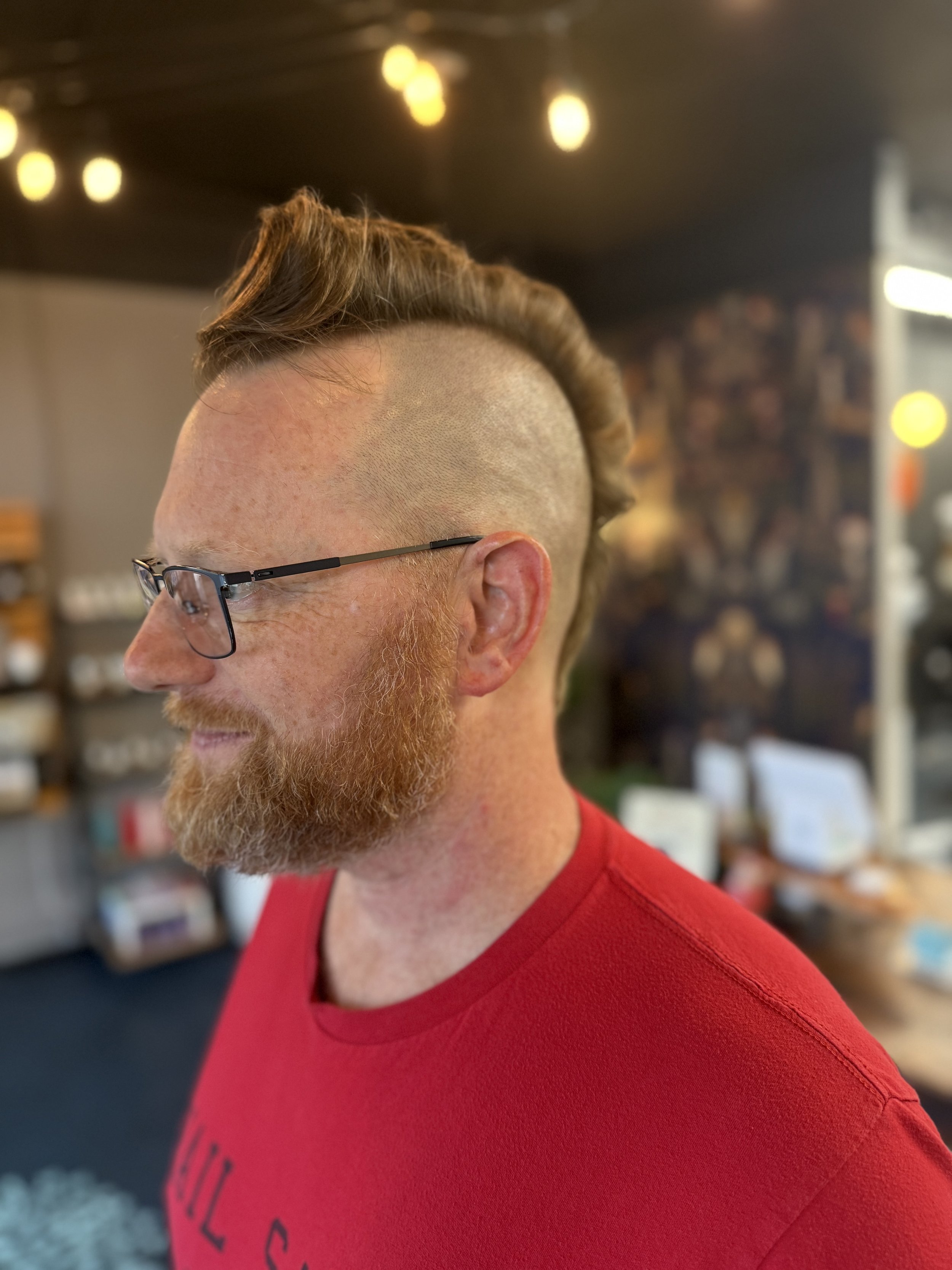 Side profile of a man with a mohawk, wearing glasses and a red shirt in a barbershop.