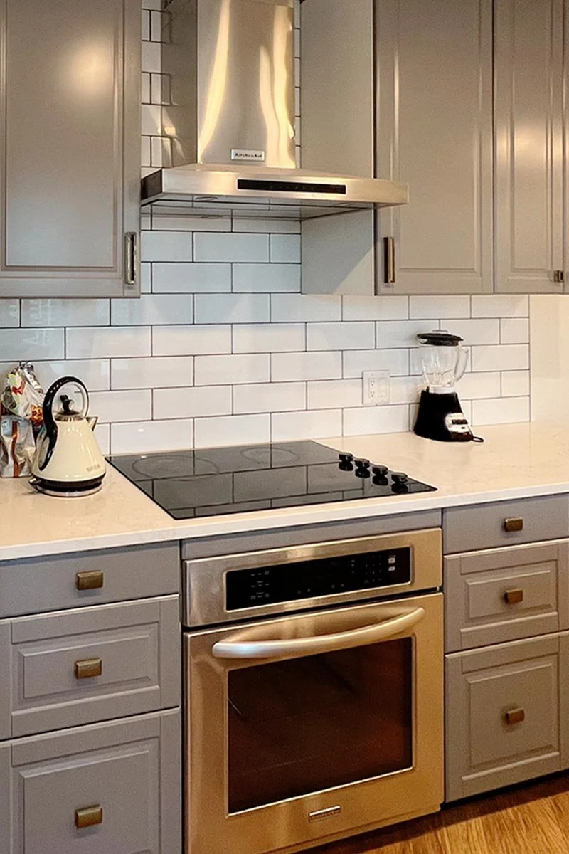Kitchen with gray cabinets, white subway tile backsplash, stainless steel oven and range hood, black glass cooktop, kettle, blender, and countertop.