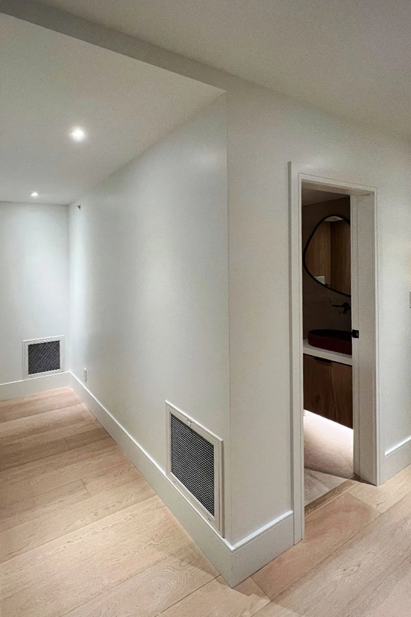 A corner of a modern interior with white walls, wooden flooring, recessed lighting, and a small doorway leading to a bathroom with a circular mirror and a vessel sink.