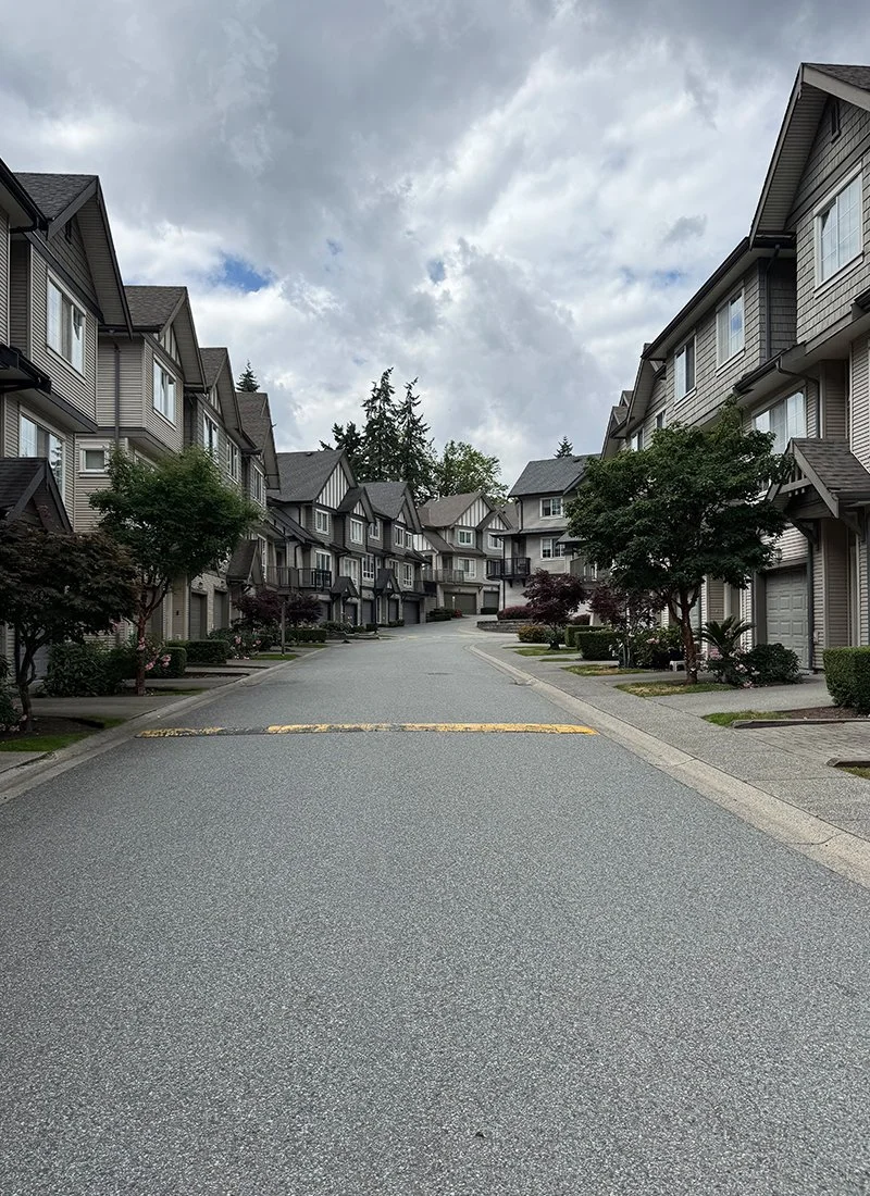 Residential street lined with multi-story houses and trees, under cloudy sky.