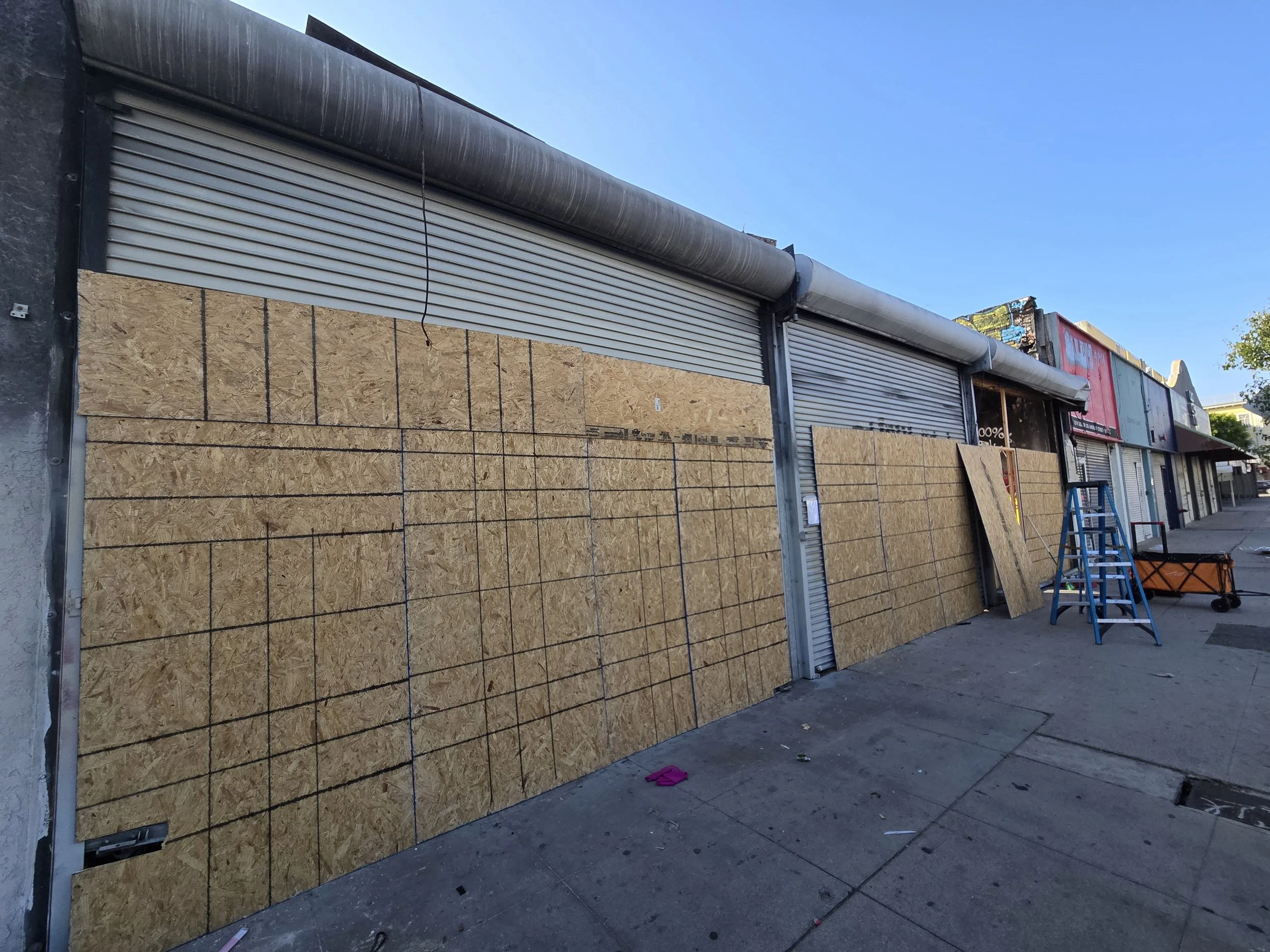 A long commercial street showing multiple businesses with large plywood board-ups and corrugated metal security shutters.