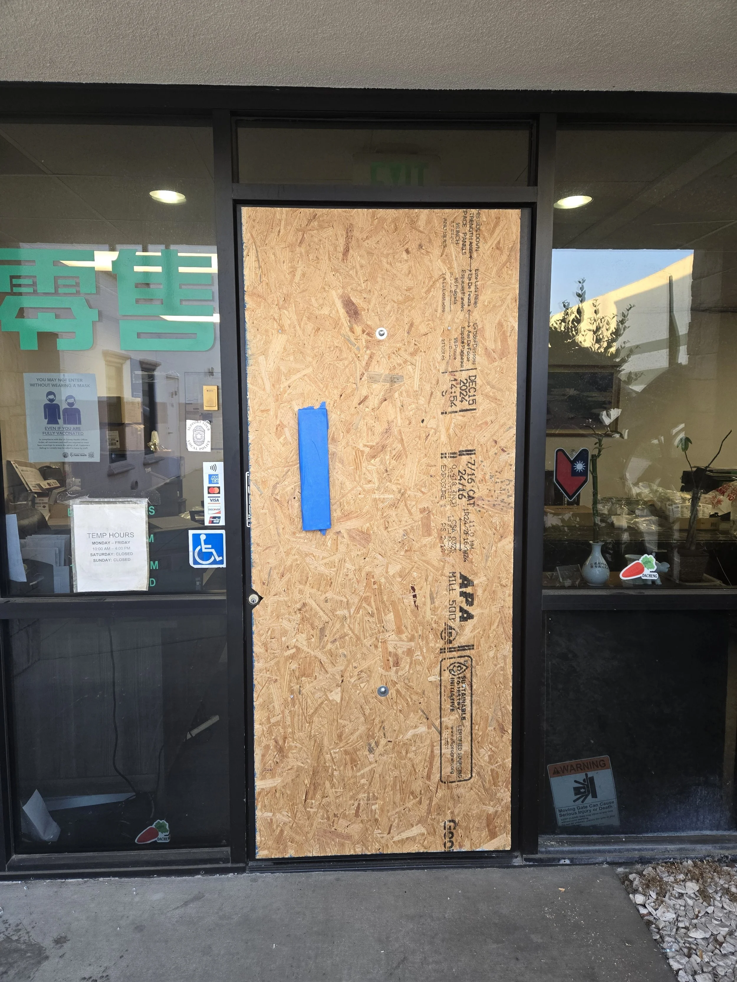 Exterior view of a business entrance featuring a temporary board-up with an OSB panel, set within a glass facade.