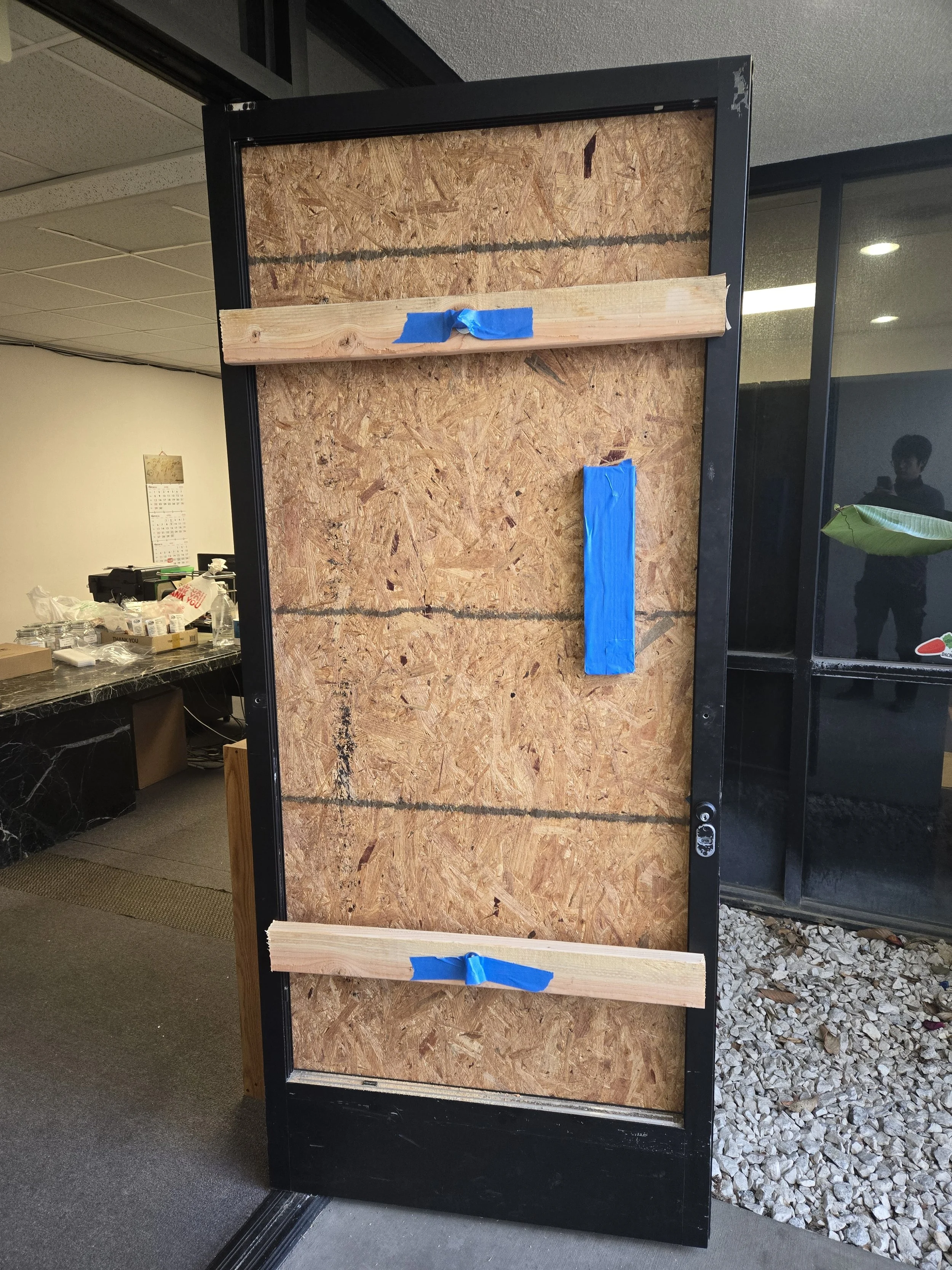 Close-up of a temporary board-up in a doorway, featuring an OSB panel with two visible wooden cross-supports and blue tape.