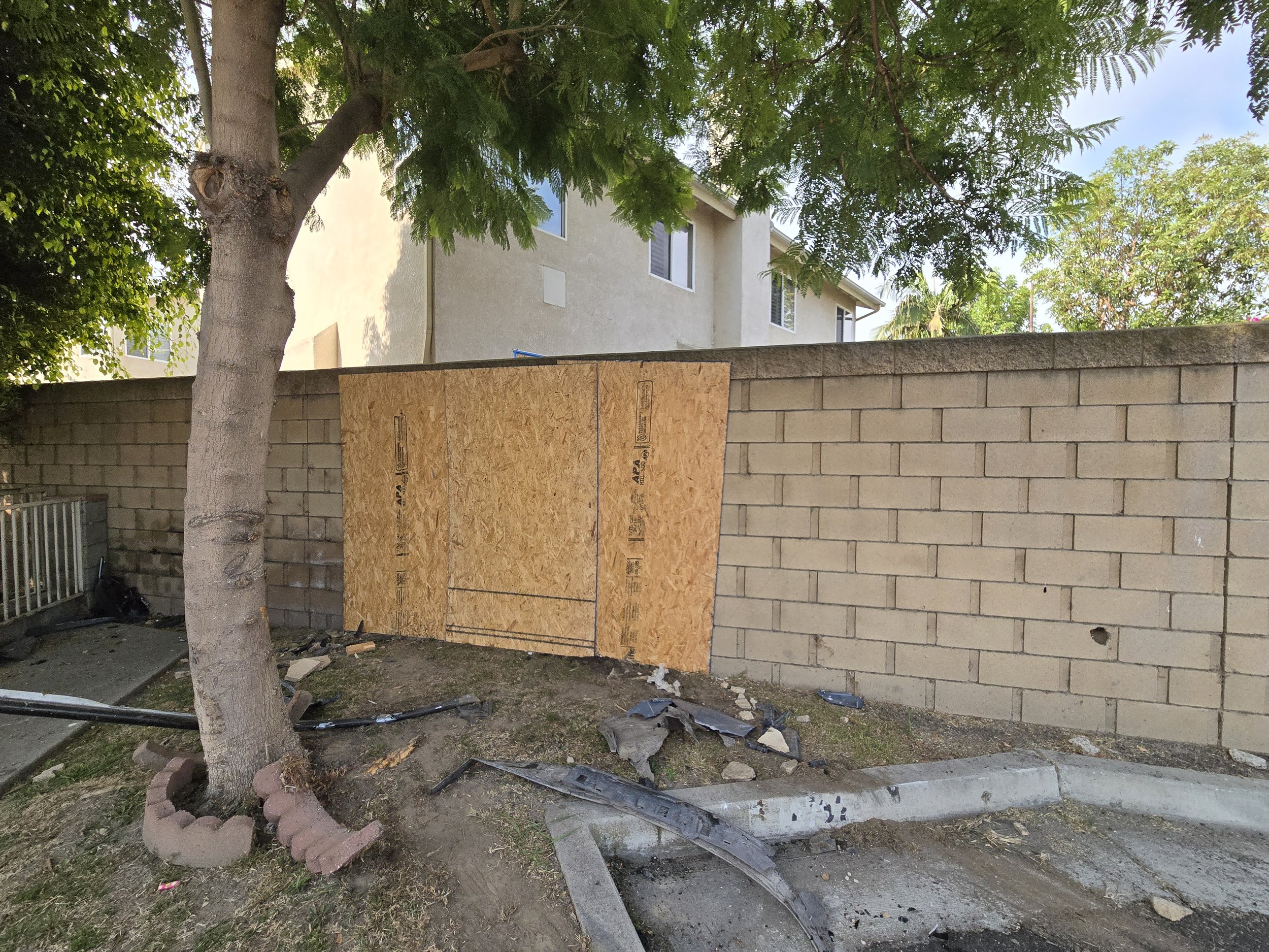 
An outdoor daytime photo shows a yard with a concrete block wall on the right and a residential building in the background. A section of the block wall has been replaced or covered with a large sheet of plywood, installed to secure or repair the are