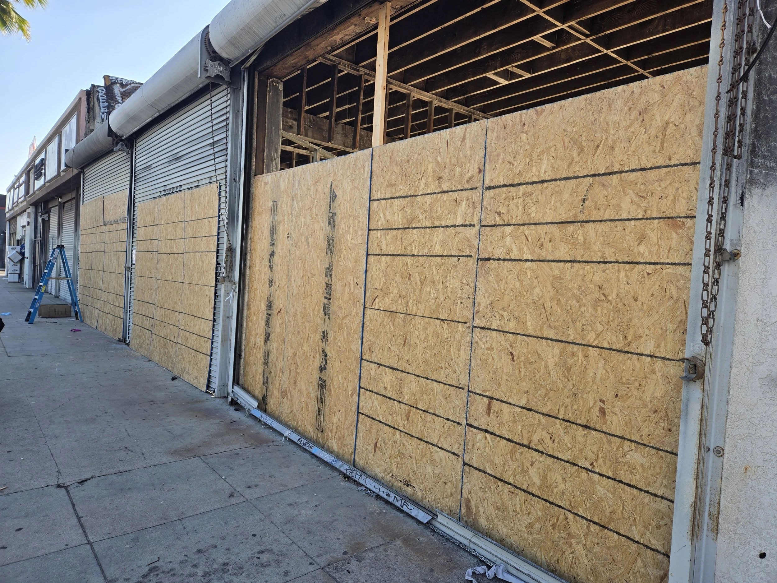 Exterior of a building showing extensive temporary board-ups with plywood, alongside closed metal rolling security gates.