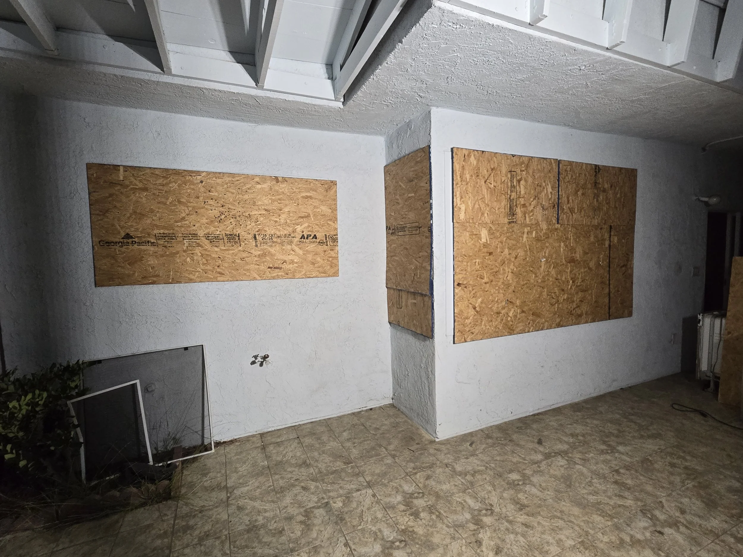 
An indoor view of a corner of a room with two large windows boarded up with plywood sheets. The walls are a light color and the ceiling has exposed beams painted white. The floor is covered with a patterned linoleum or tile, and a screen door and an