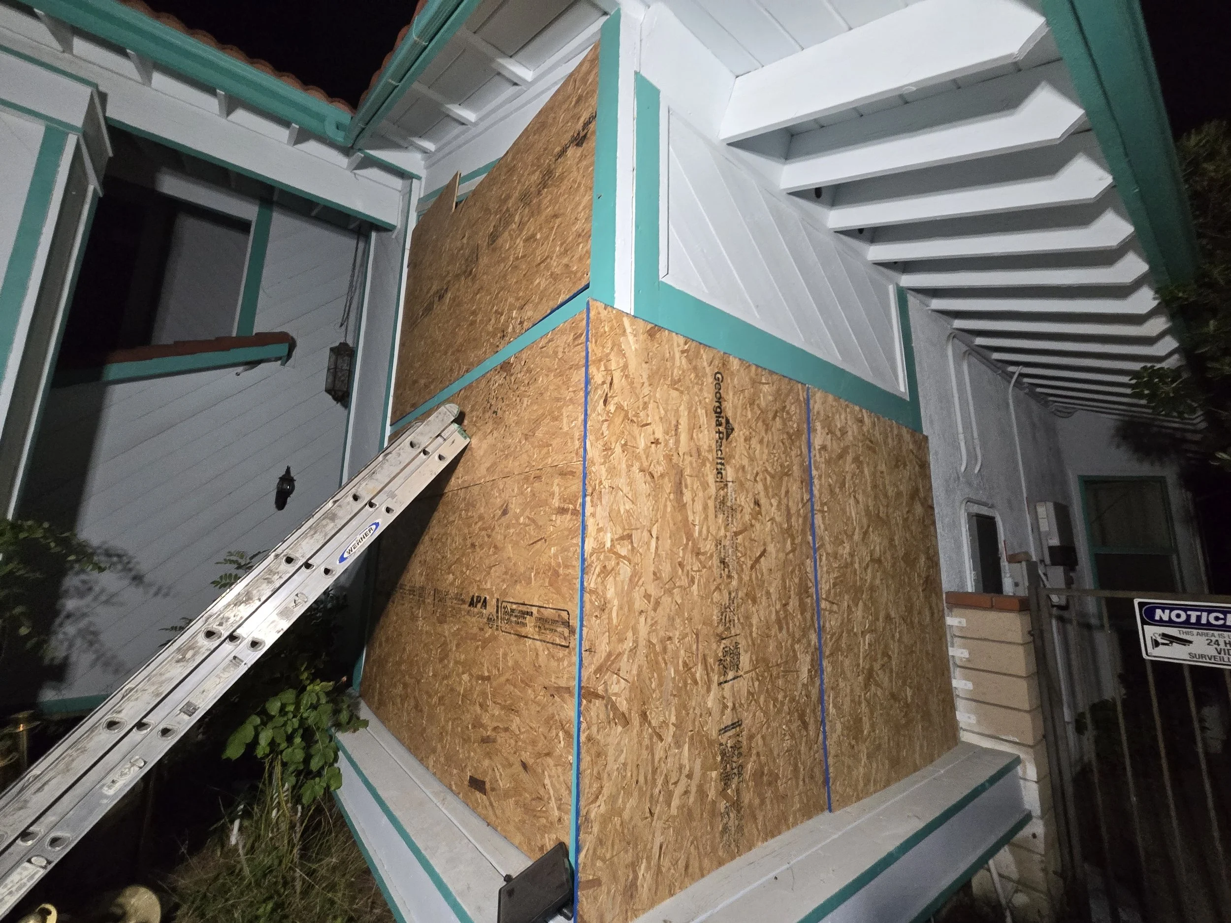 
Two exterior walls of a house, one with a window and the other without, are boarded up with large sheets of plywood. The house siding is painted white with bright teal trim. An extension ladder is propped up against the house on the left side of the