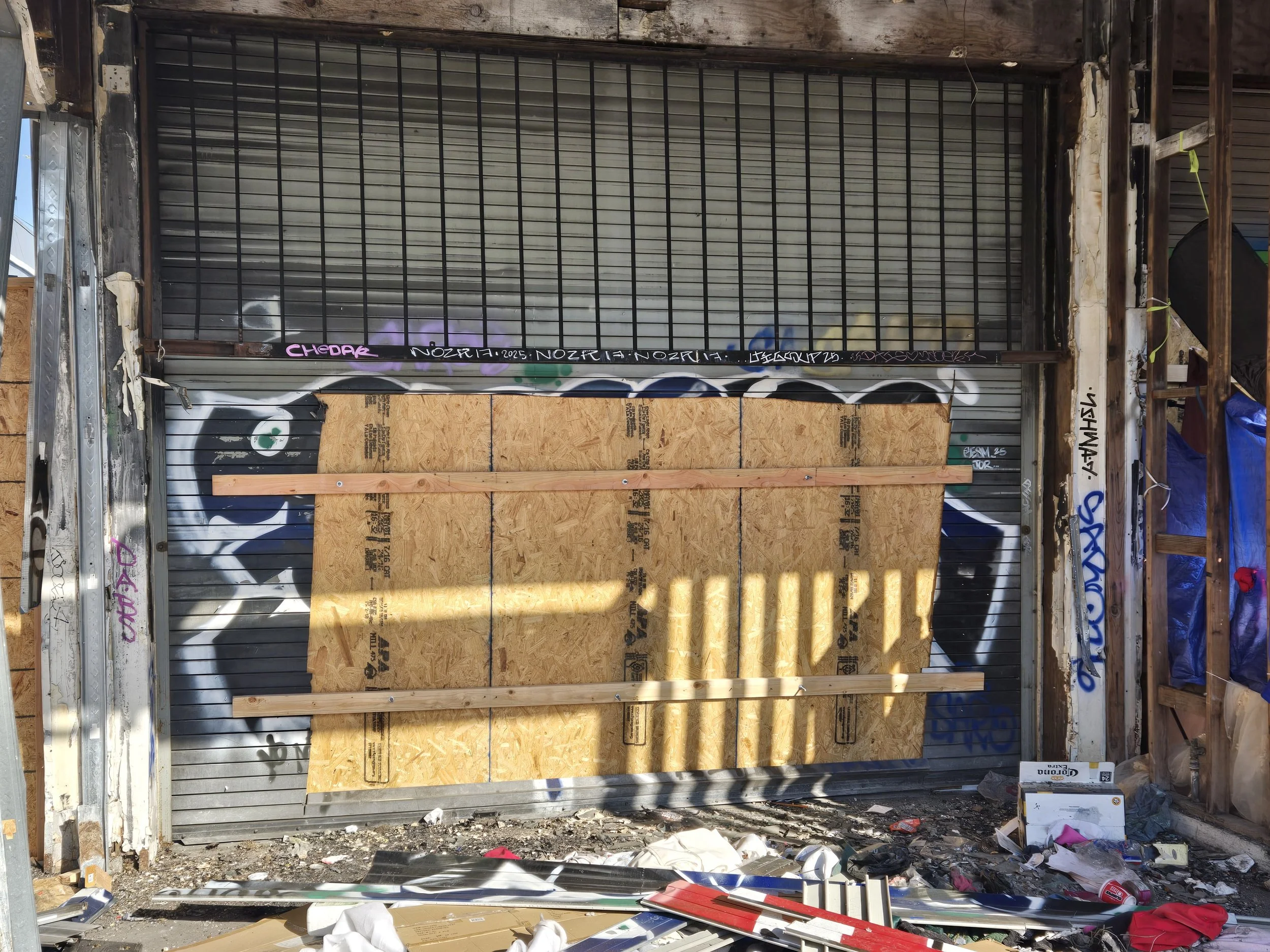 A wide temporary board-up made of plywood with horizontal wooden braces, set against a backdrop of graffitied metal shutters.