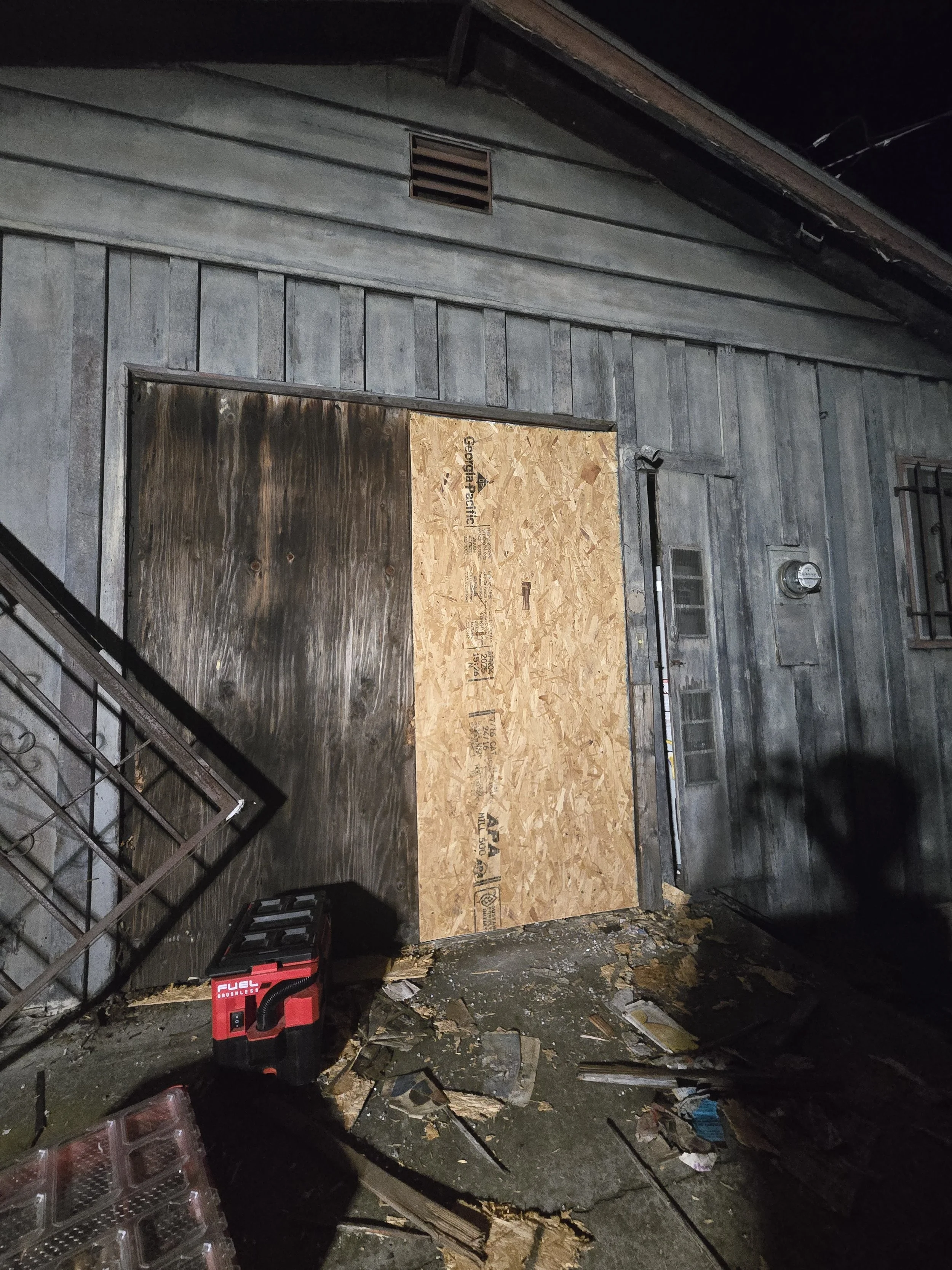 Residential door board-up in Los Angeles — plywood securing a 
damaged entrance on an older wood-sided home. Late-night emergency 
response by Boardup LA.