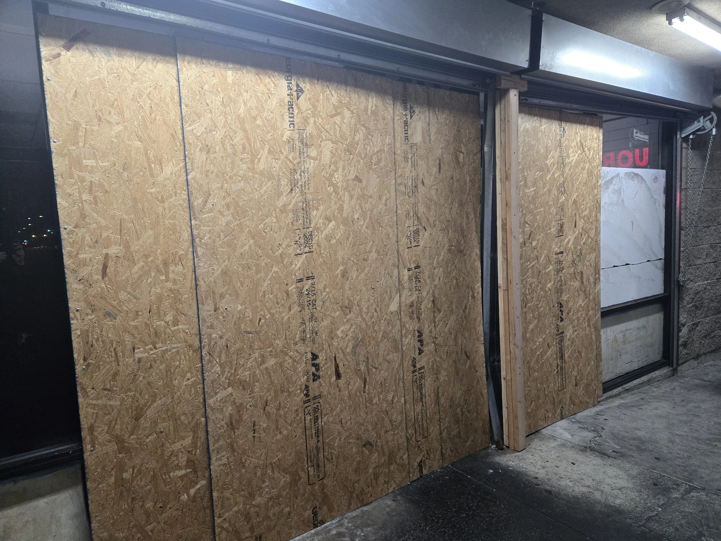 Close-up of a sturdy board-up: a large OSB panel filling an entrance, with a strong wooden support beam visible.