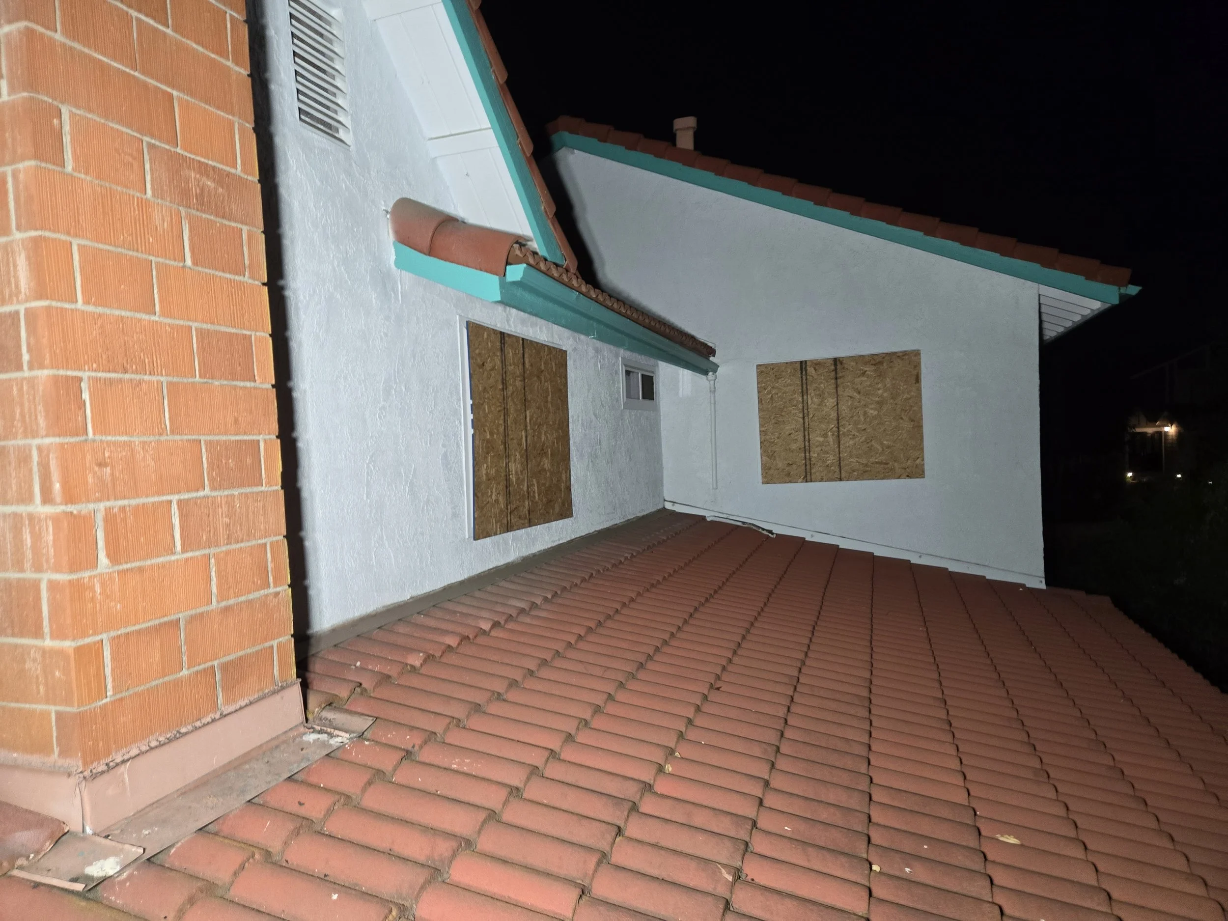 
An elevated, outdoor view of a building at night, showing a roof with terracotta-colored tiles. A brick chimney is visible on the left side of the image. Two windows on the upper portion of the building's wall are boarded up with plywood sheets. The