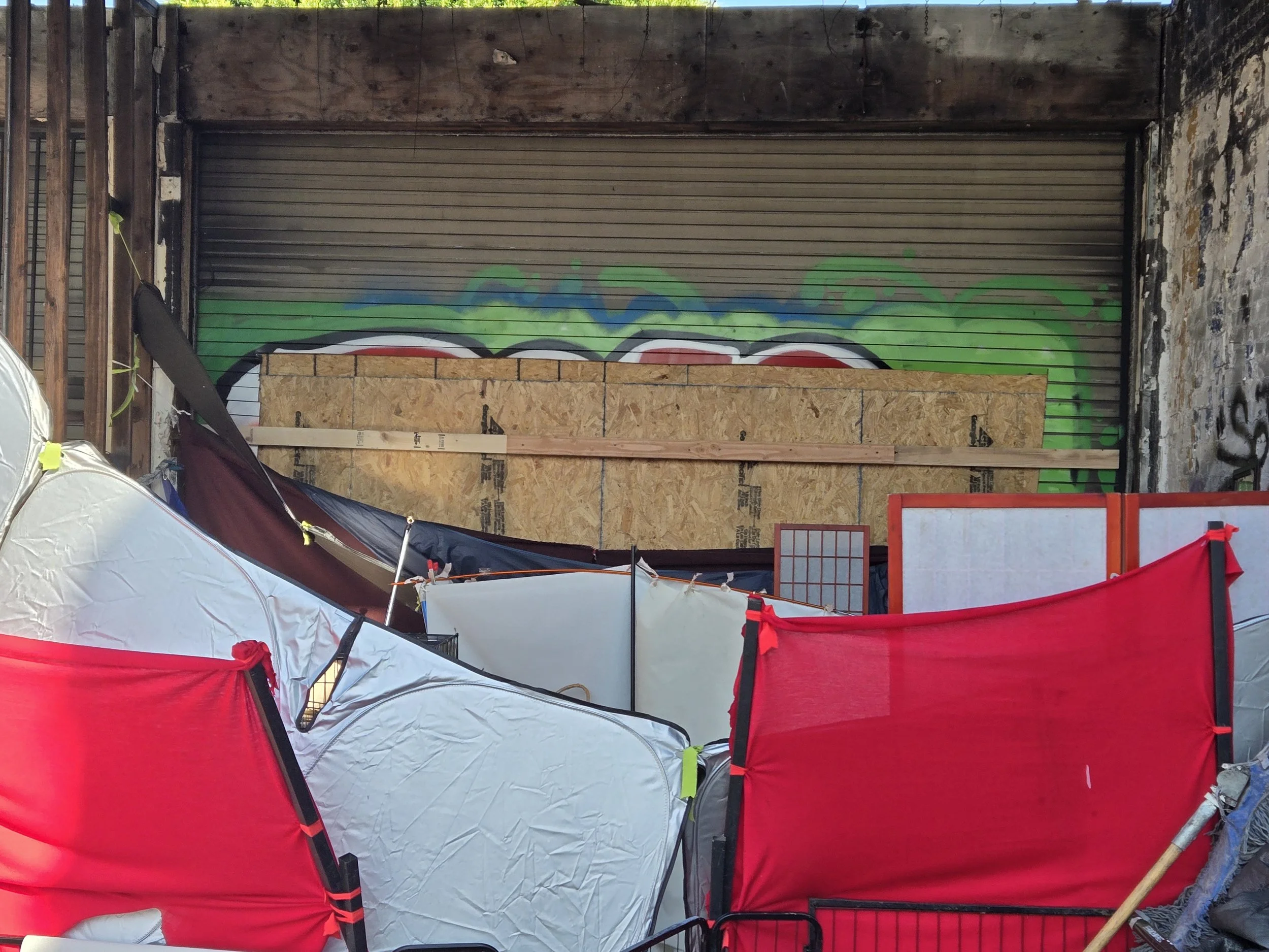 A rough interior setting with a temporary plywood wall, graffiti, and various items including red and silver coverings.