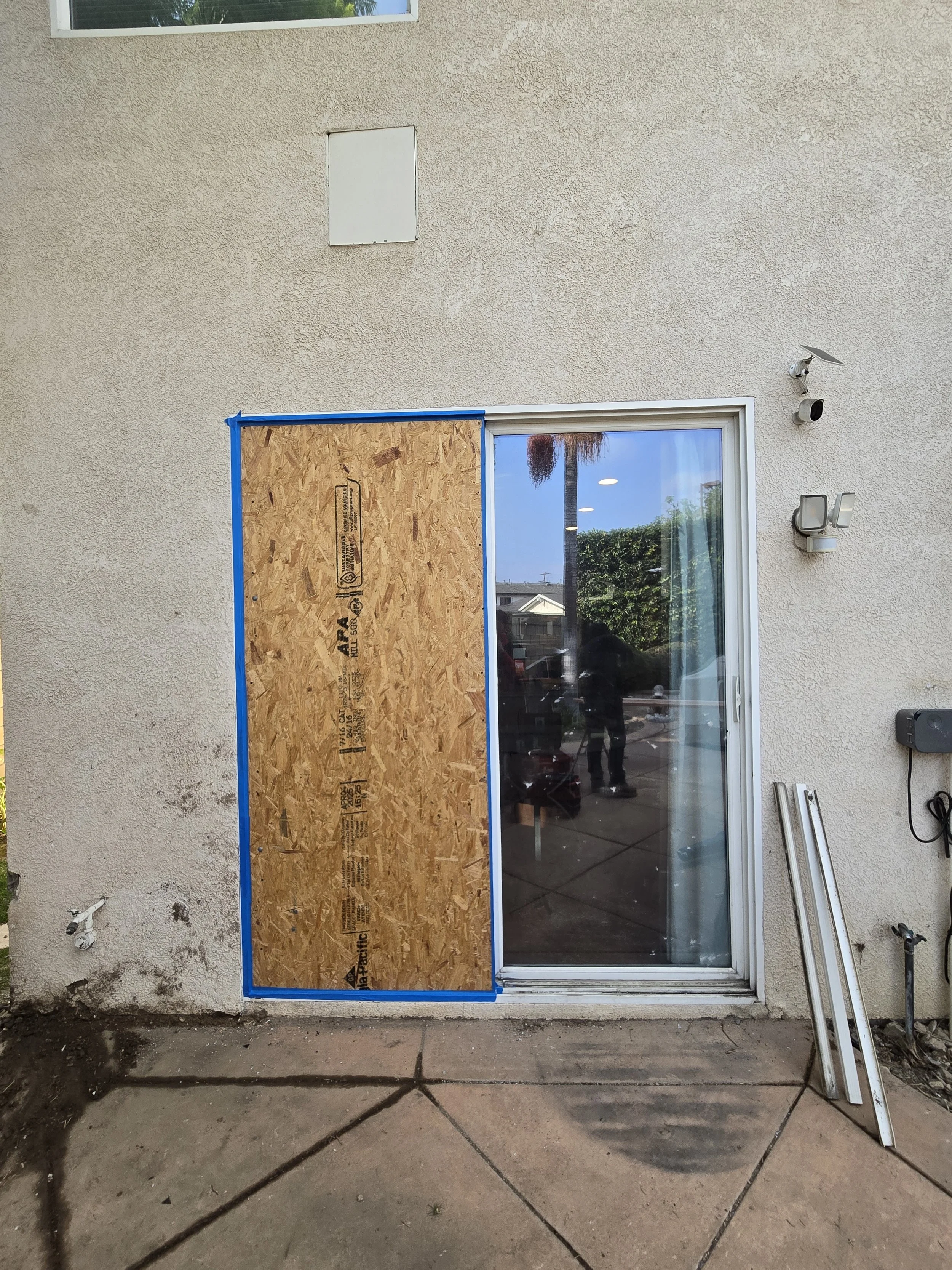 
An outdoor daytime photo of a sliding glass door on the exterior of a home. One of the glass panels has been boarded up with a sheet of plywood, secured with what appears to be blue tape around the edges to protect the remaining glass and frame. The