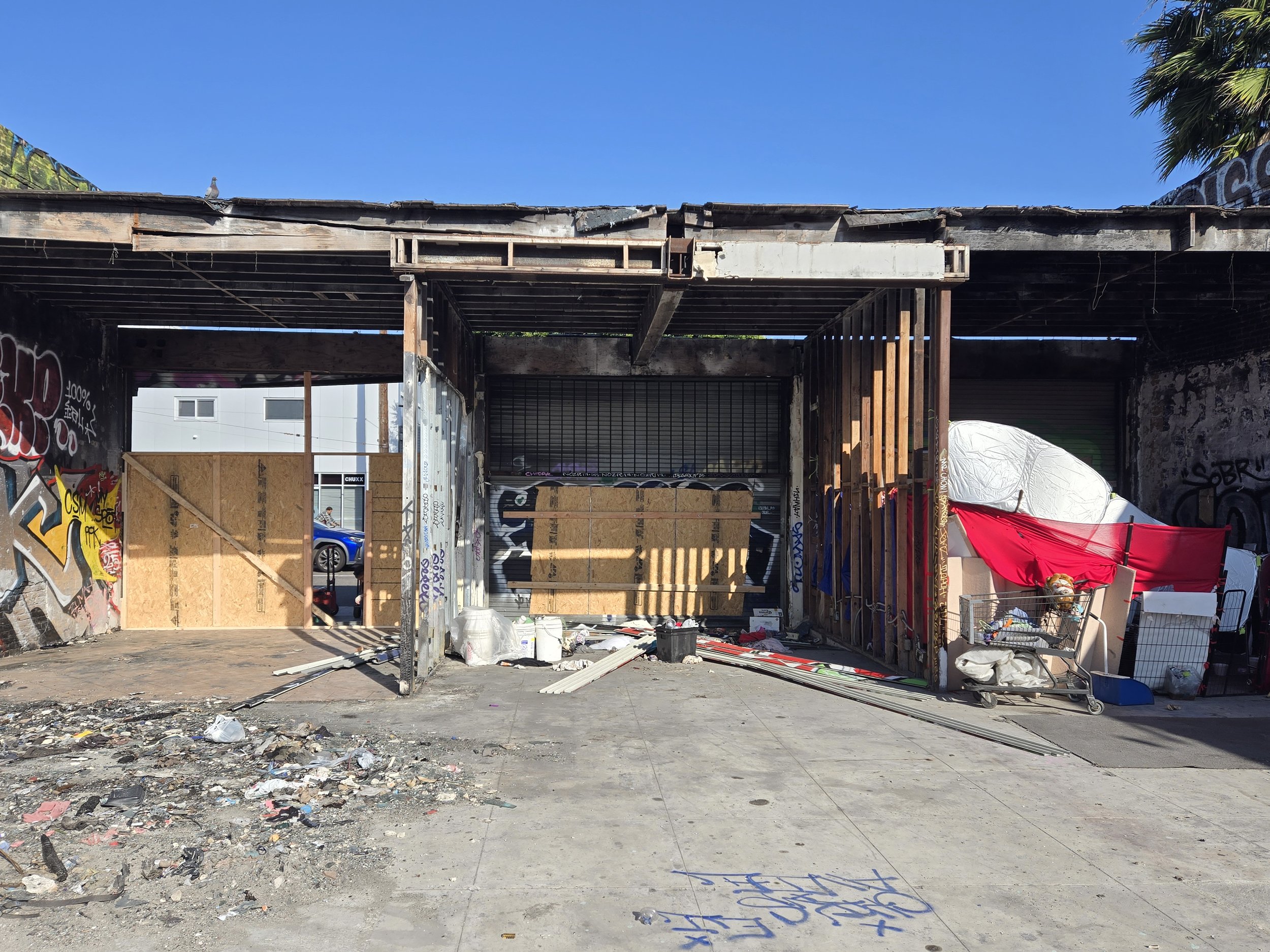 Exterior of a heavily damaged building with exposed framework, debris, graffiti, and some temporary plywood boarding.