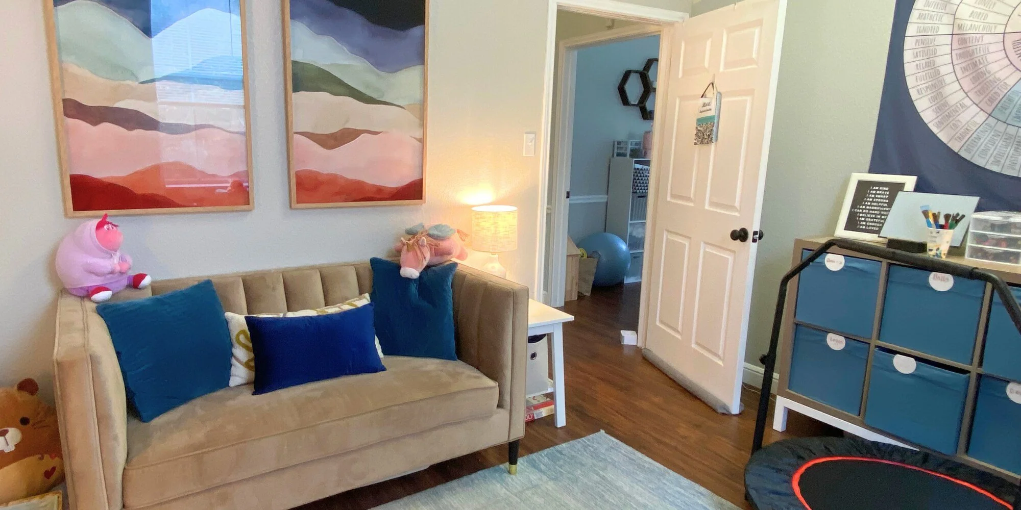 Child's playroom with beige sofa, blue pillows, plush toys, wall art, side table with lamp, and a storage unit with labeled bins.