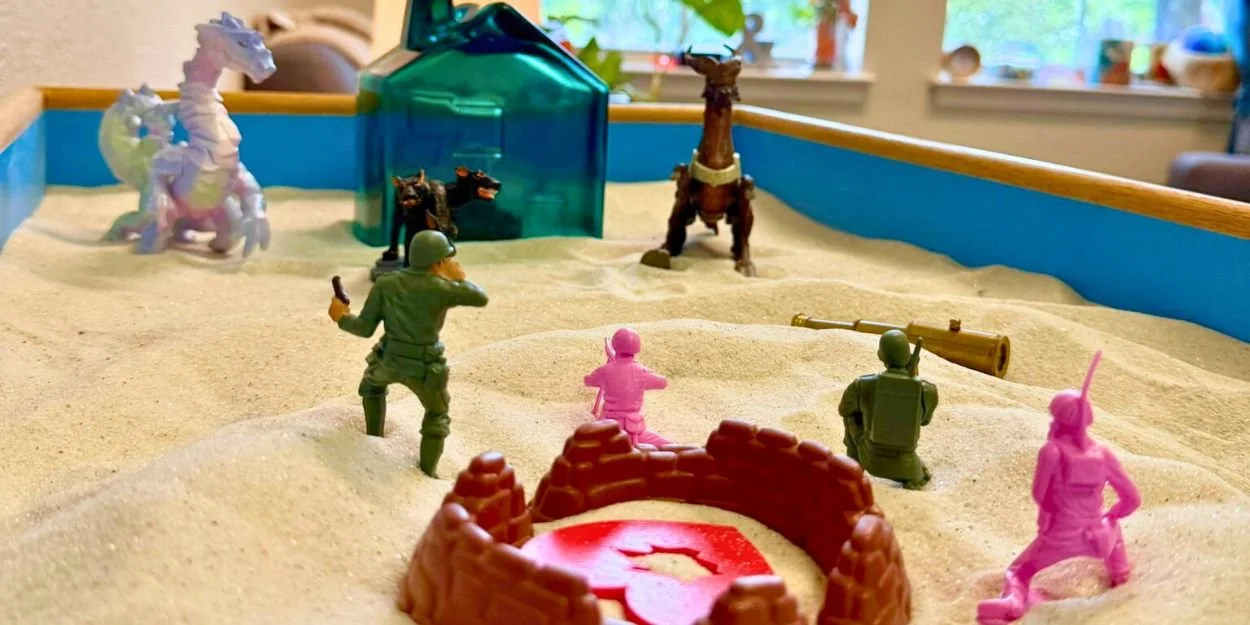 Toy soldiers and animals in a sandbox with sand dunes, a transparent blue plastic house, and toy cannons.