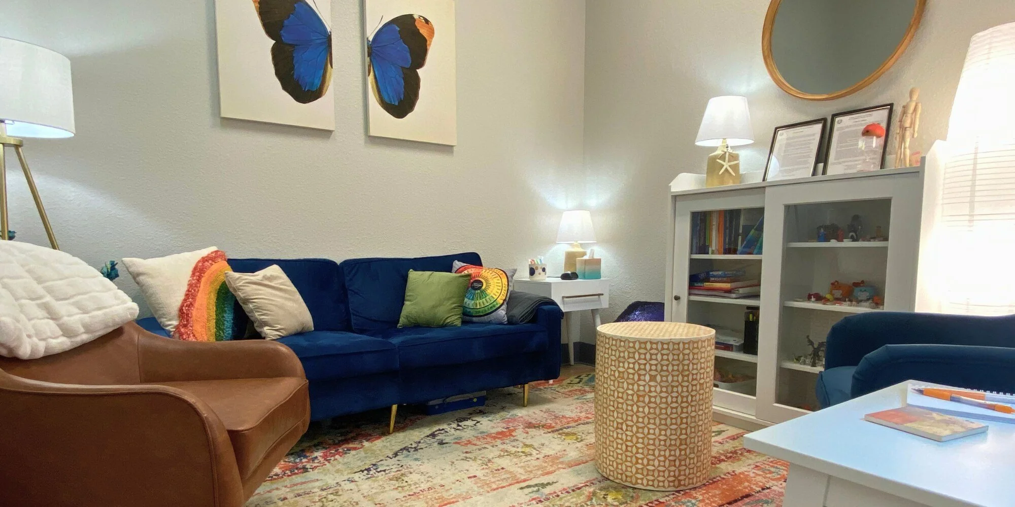 Living room with a blue velvet sofa decorated with multicolored throw pillows, a brown leather armchair, a white side table with a lamp, a white bookcase with framed pictures on top, a colorful rug, and wall art depicting a butterfly.